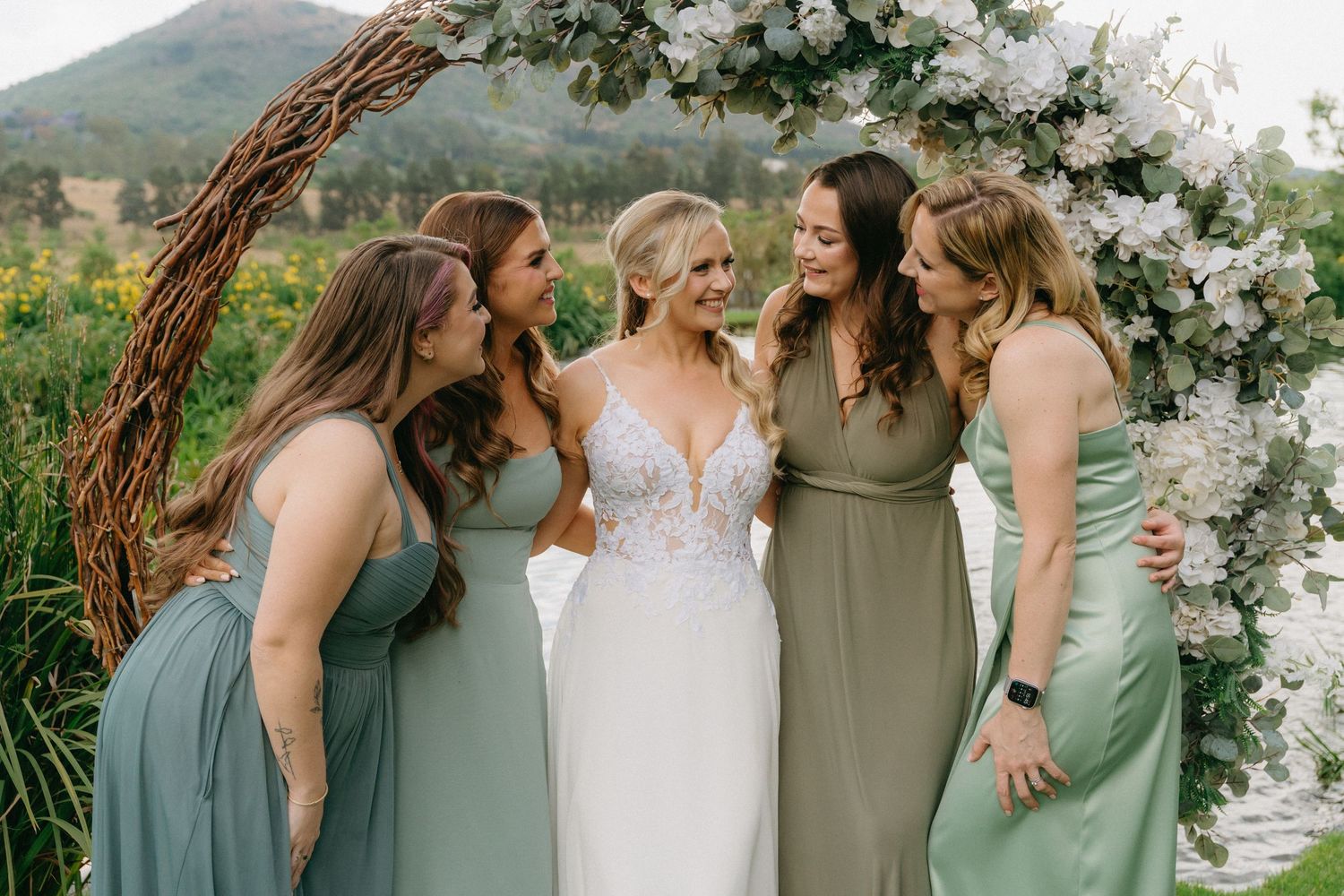 Bridesmaids in sage green dresses gather under a floral arch outdoors.