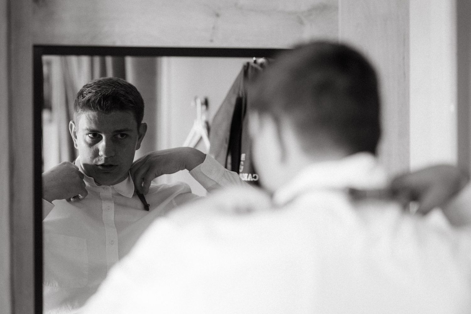A black and white reflection in a mirror shows someone adjusting a white dress shirt collar at home.
