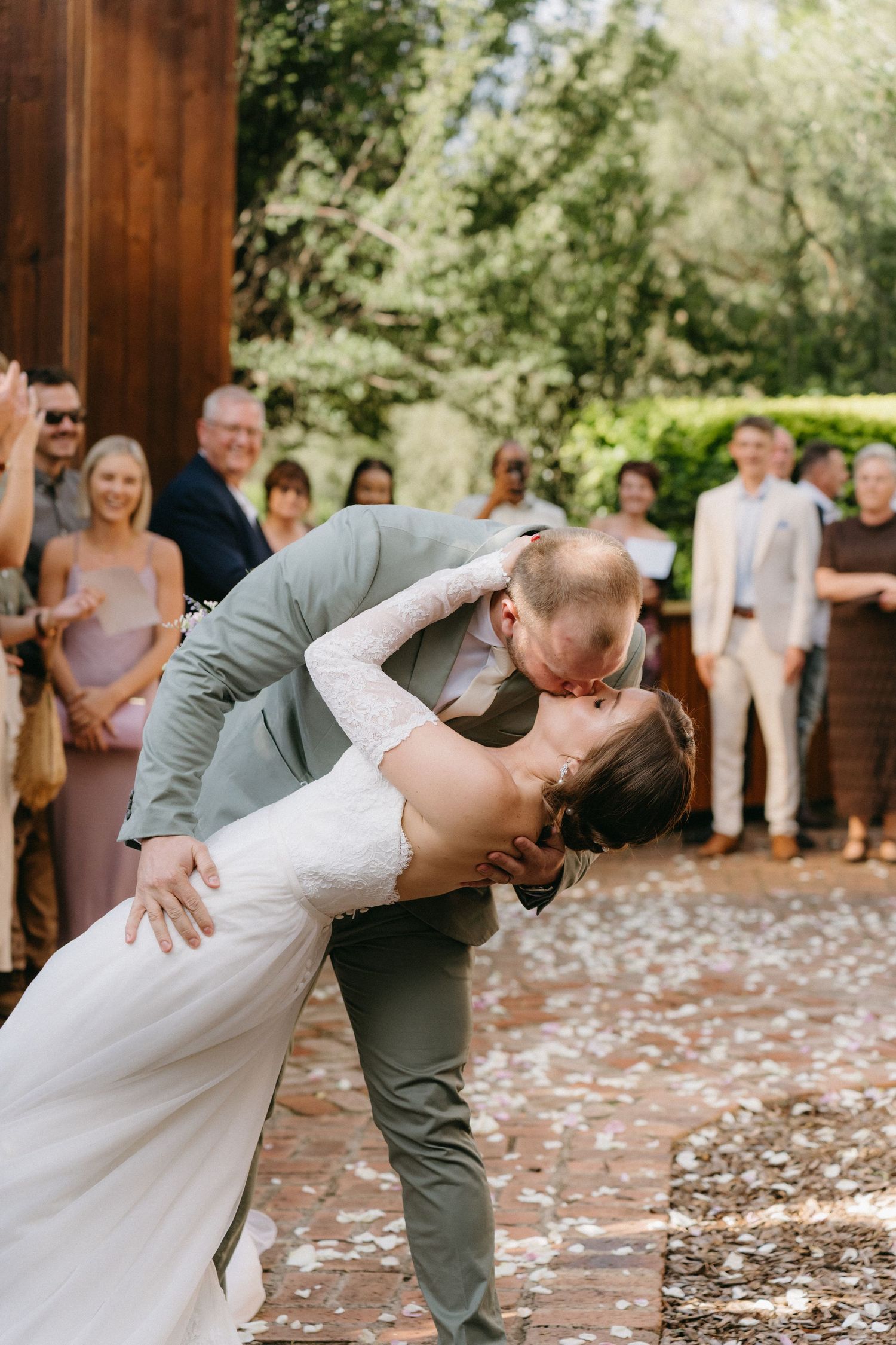 Romantic wedding couple sharing a dip kiss outdoors with guests watching in the background.