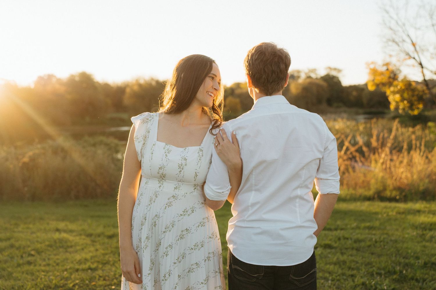 A couple standing in a golden sunlit meadow at sunset as rays illuminate their silhouettes from behind.