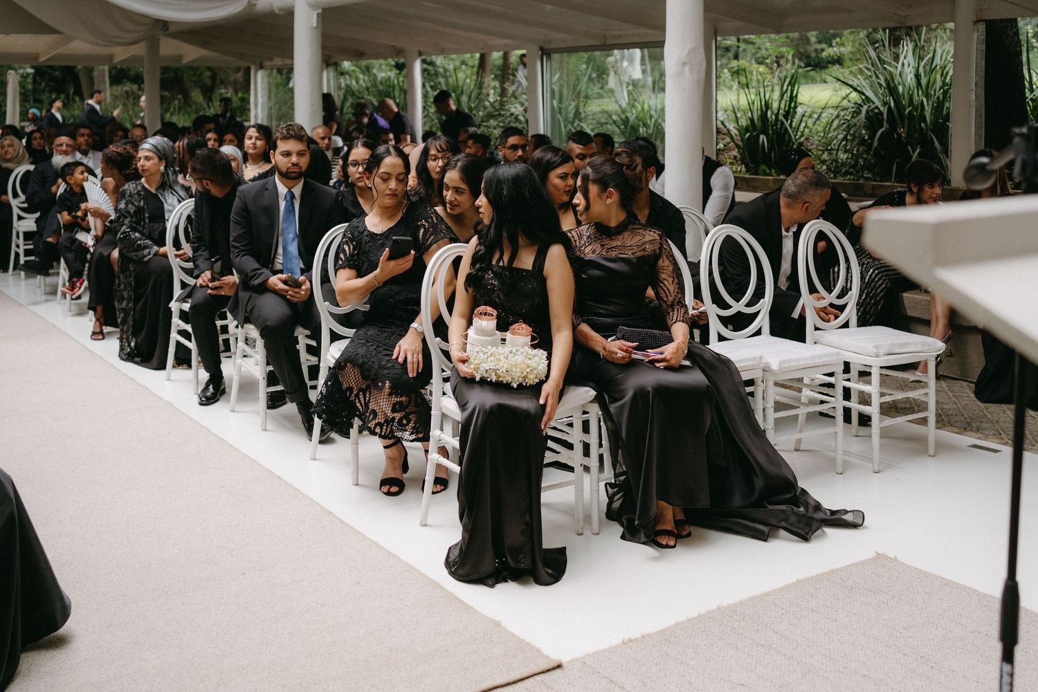 Wedding guests in formal black attire seated on white chairs at an elegant outdoor ceremony venue.