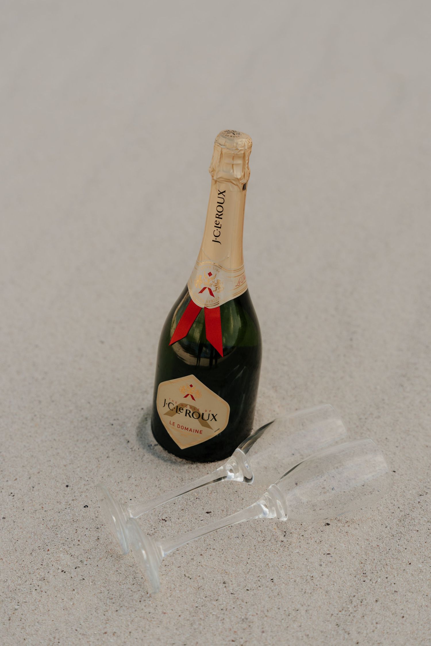 J.Caproux champagne bottle with red ribbon and two crystal flutes lying on white sand beach.