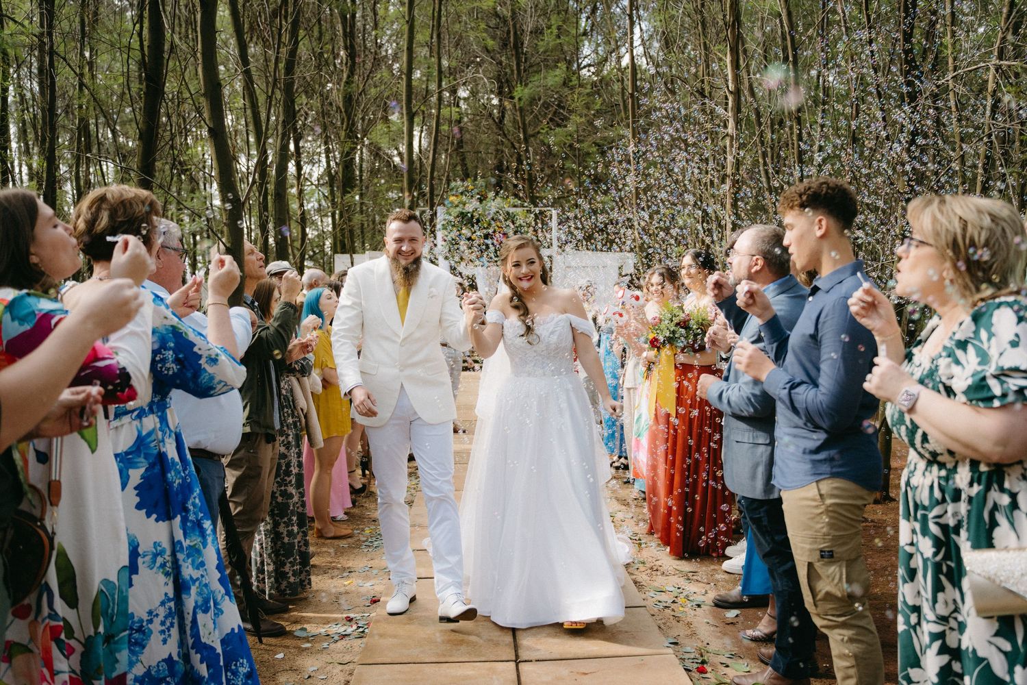 Wedding guests line a wooden walkway through the woods celebrating the newlyweds.