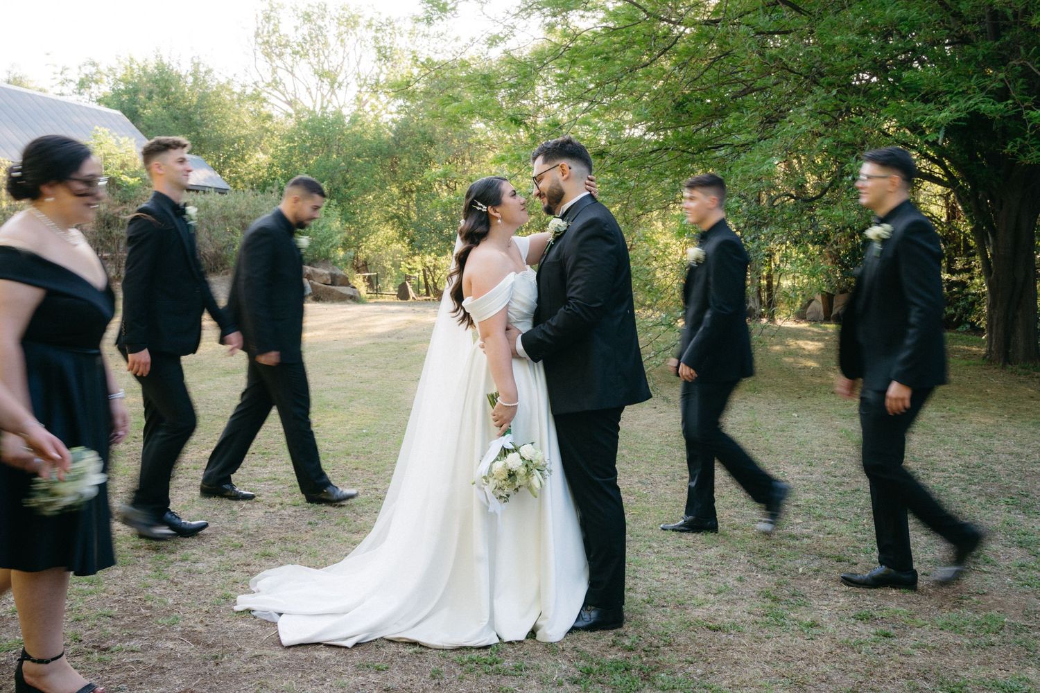 Wedding party in black attire walks through outdoor garden setting as newlywed couple embraces in center.