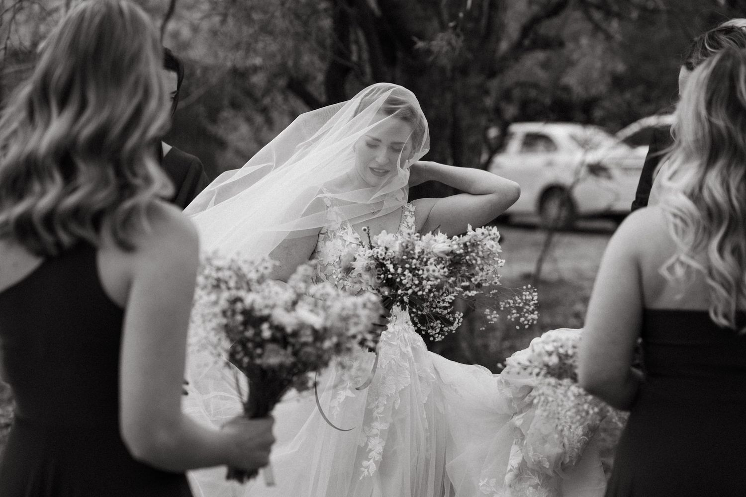 A black and white photo of bridesmaids helping with a bride's dress and flowers outdoors.