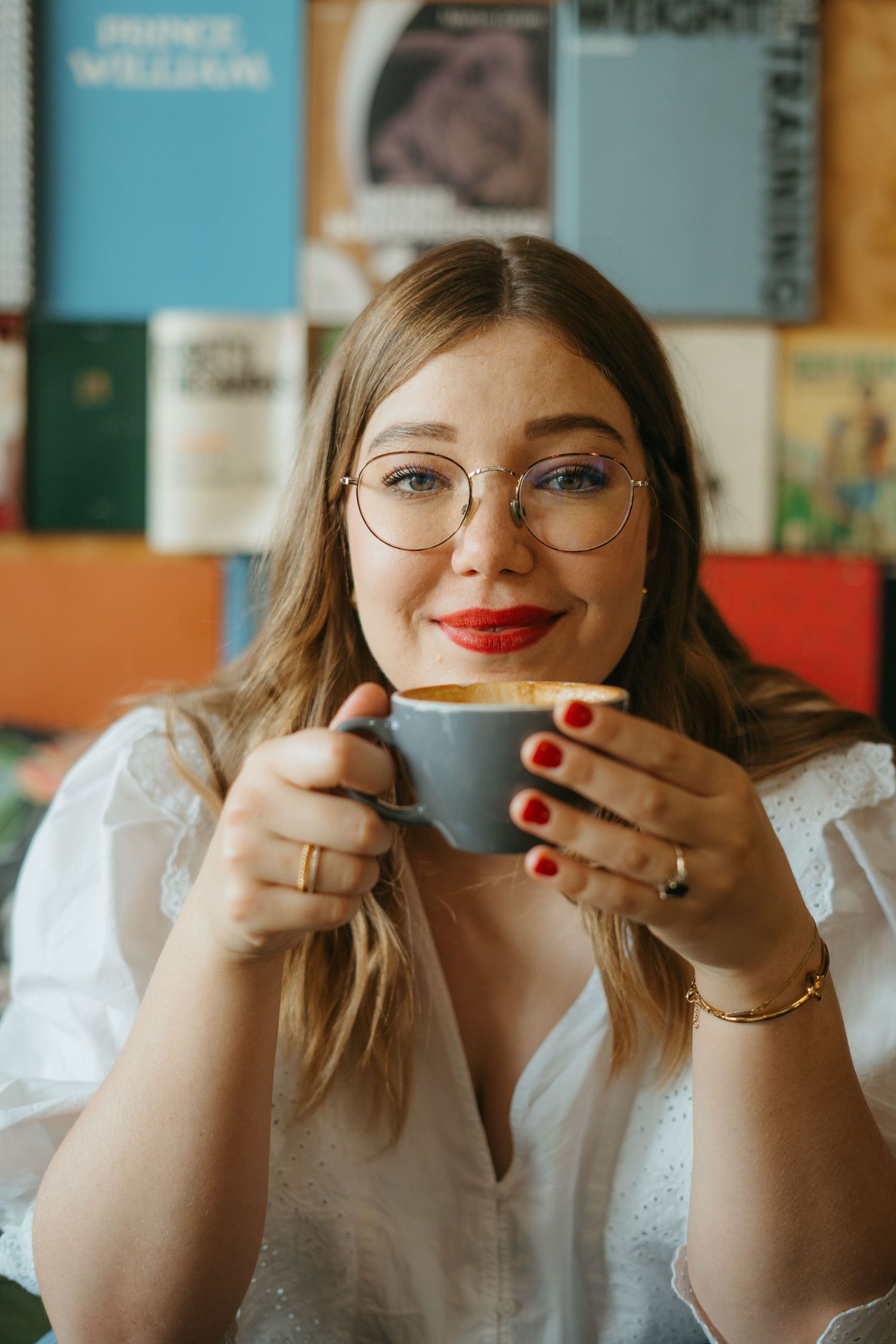 Someone holding a coffee cup wearing glasses and a white blouse in front of a bookshelf smiles while enjoying their drink.