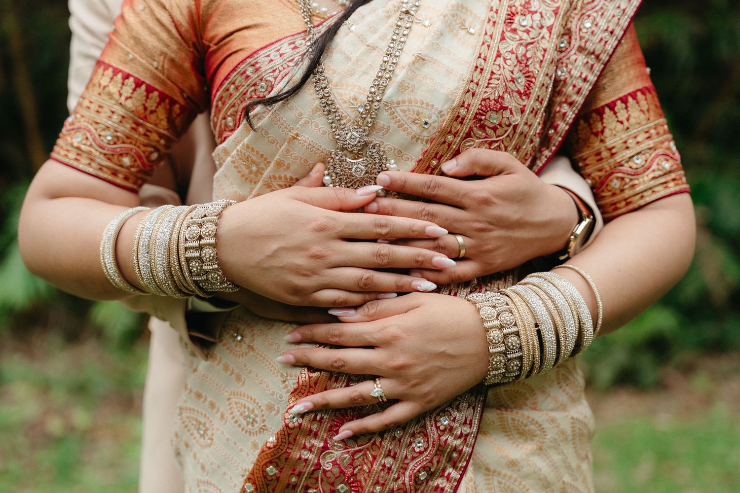 Hands adorned with traditional Indian bangles and jewelry shown crossed over an embroidered beige and red sari.