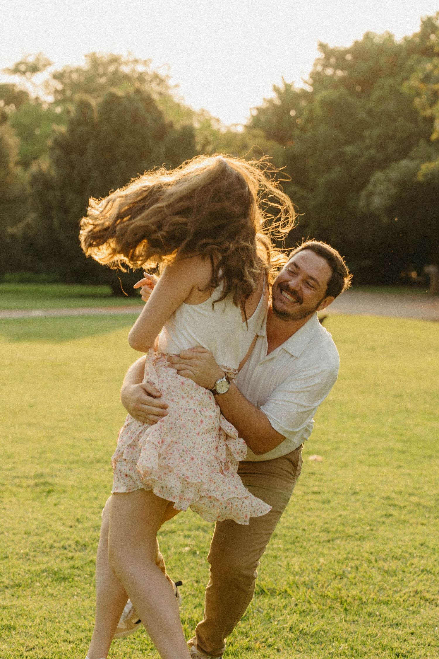 A joyful man spins a laughing woman in a sunny park, her hair flowing as they share a playful romantic moment.