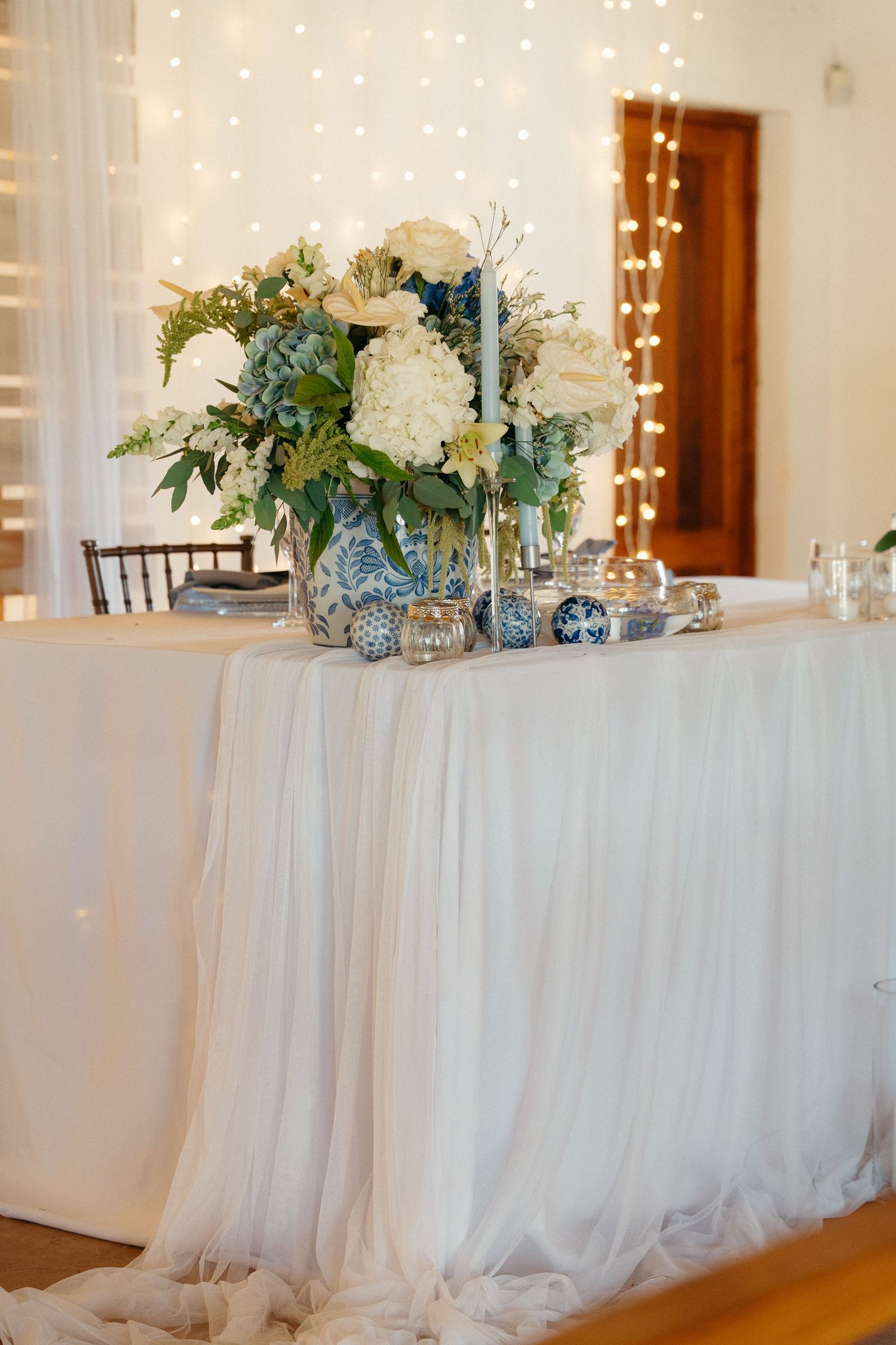 Elegant wedding reception white draped table with floral centerpieces and twinkling string lights in the background.