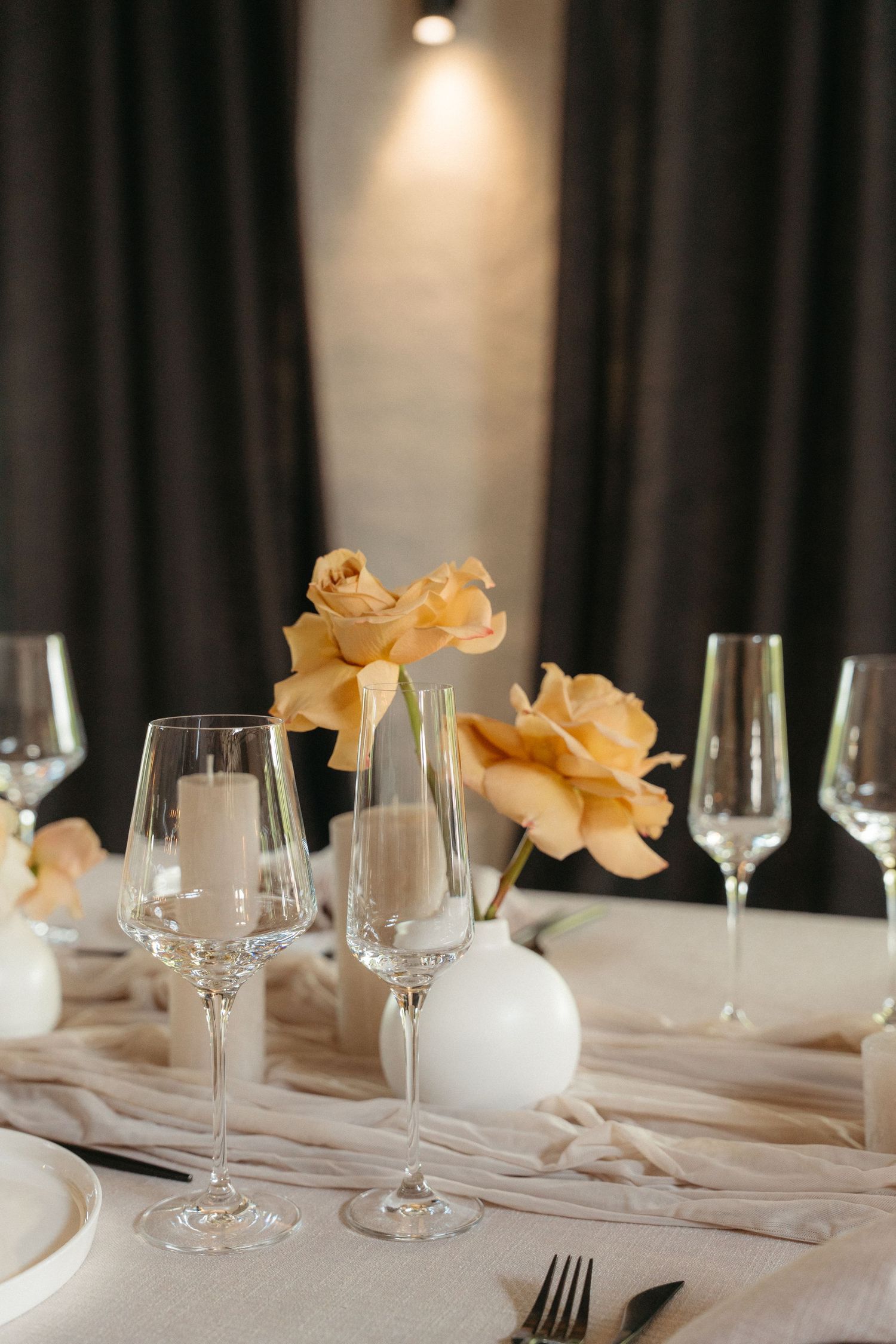 Elegant table setting with crystal wine glasses and peach roses on white tablecloth under soft lighting.