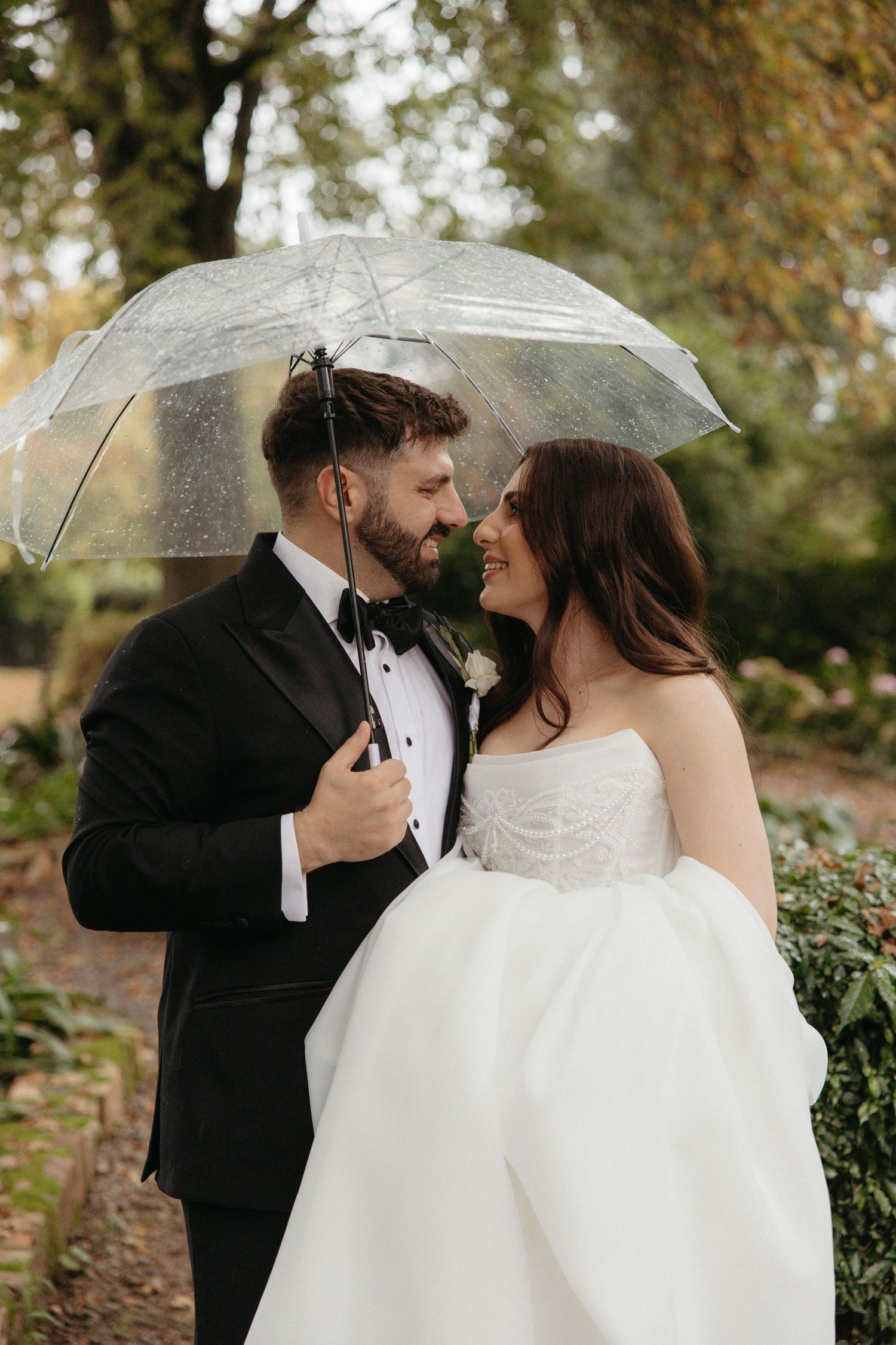 Bride and groom sharing romantic moment under clear bubble umbrella in lush garden on rainy wedding day in Johannesburg, South Africa