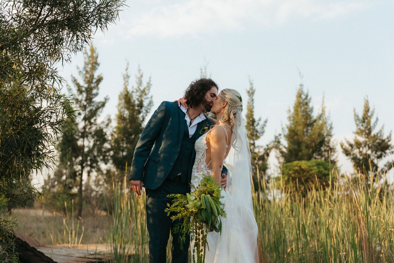 Bride and groom sharing a romantic kiss outdoors, surrounded by tall green trees and reeds on their wedding day.
