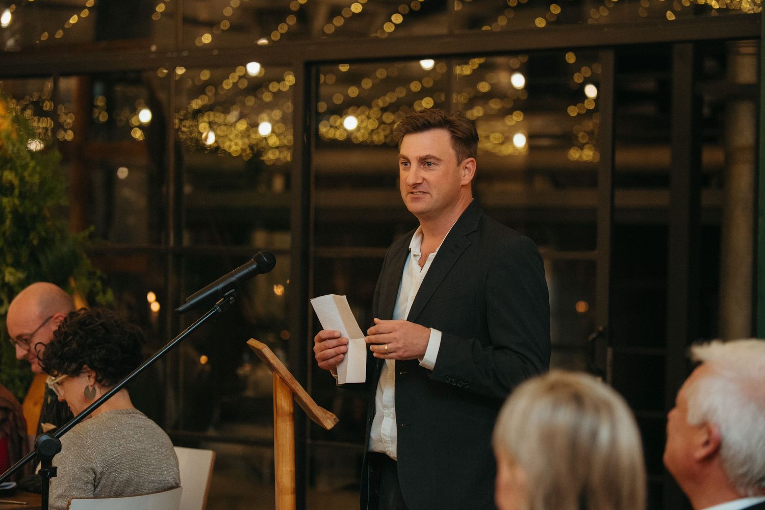 Man in dark blazer delivering speech at elegant indoor event with warm string lights in background.