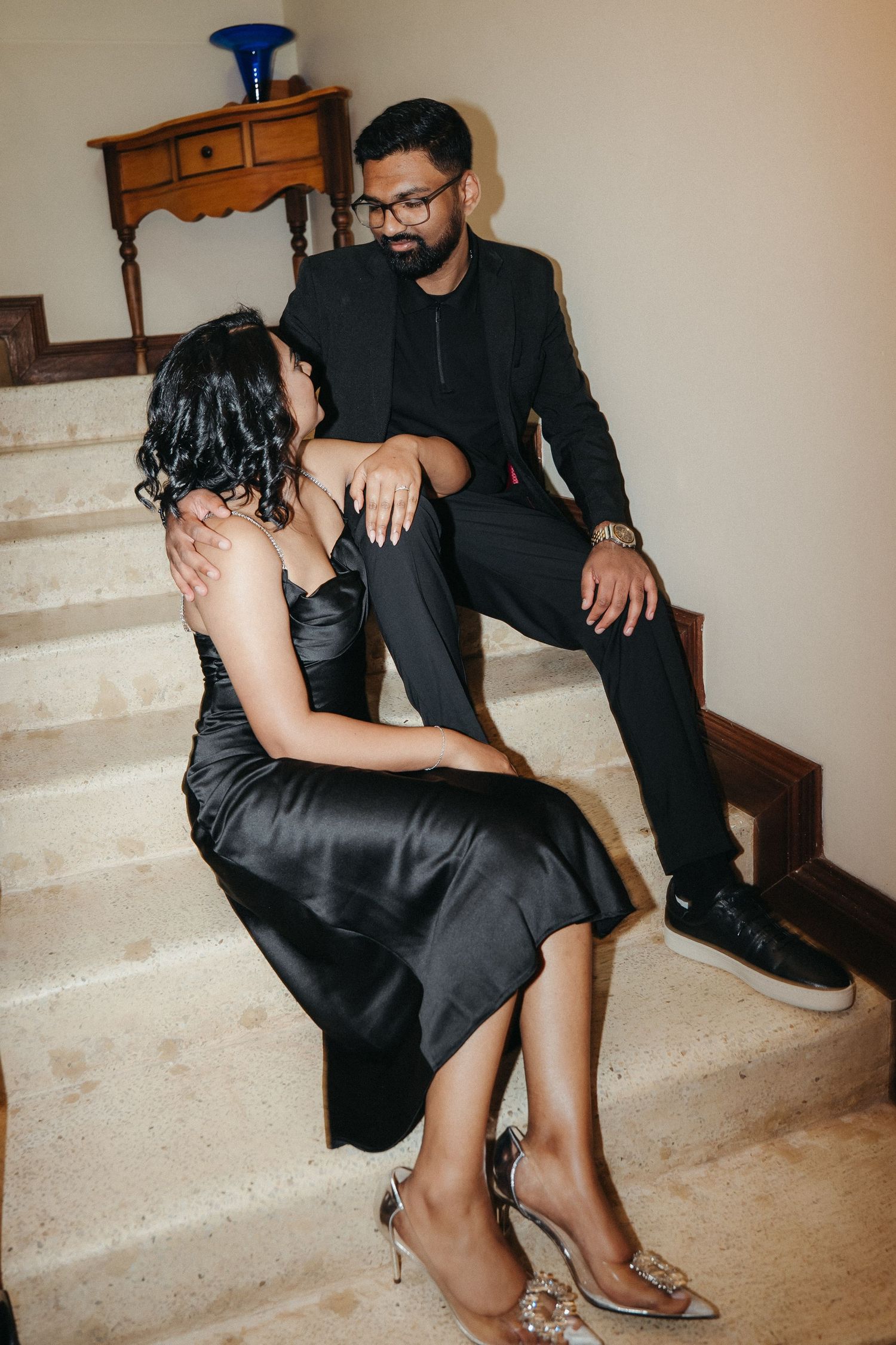 Couple in matching black outfits sitting on marble stairs, woman in satin dress with heels, man in suit with glasses.