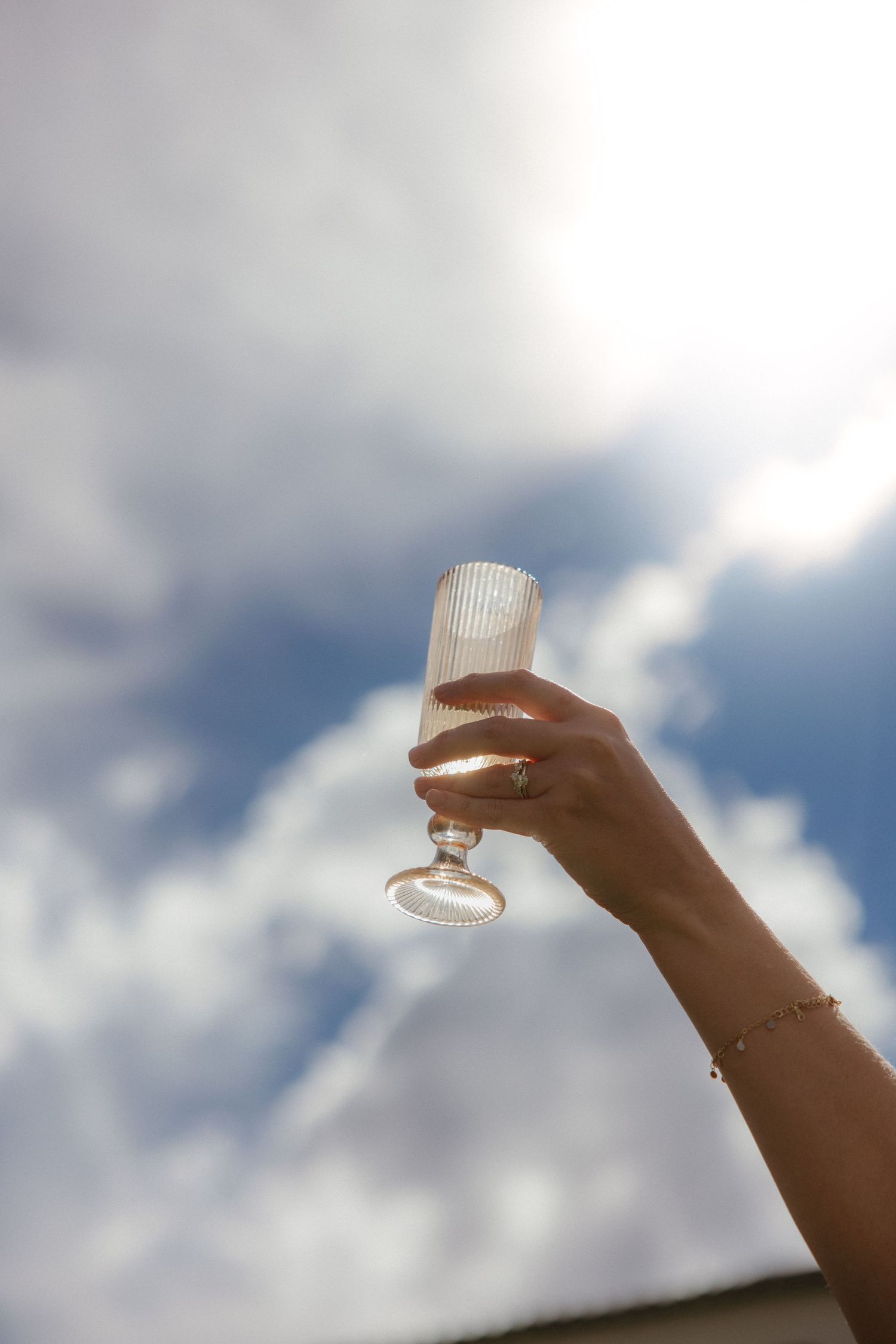 A hand holding an elegant ribbed champagne flute raised against a bright cloudy blue sky.