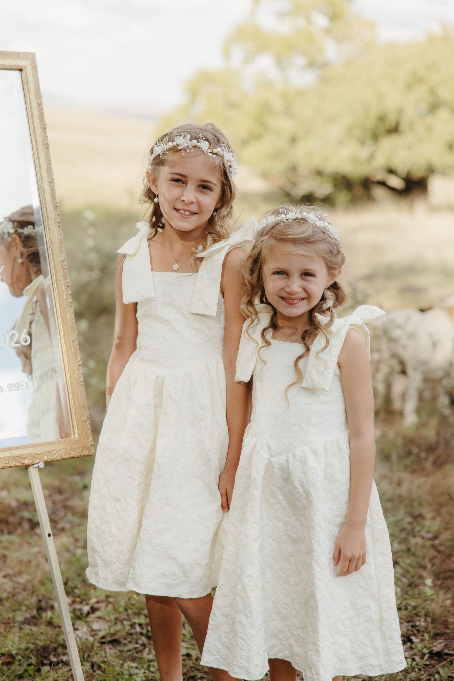 Two young flower girls in white dresses and floral headbands posing outdoors at a wedding ceremony.