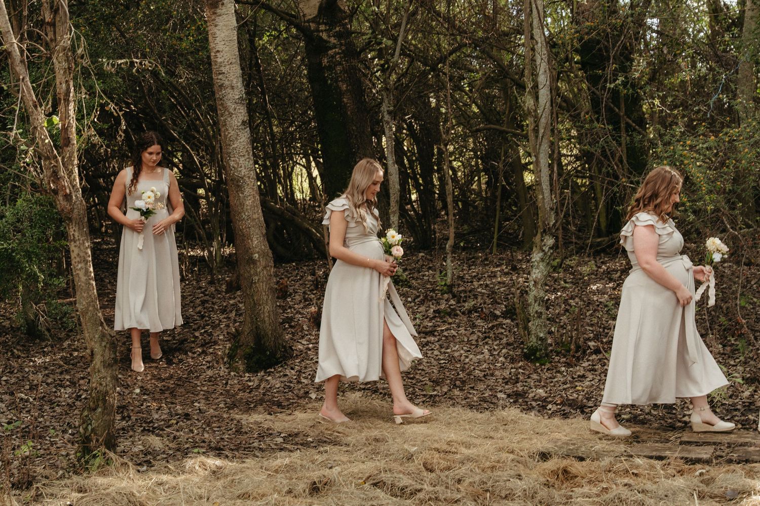 Three bridesmaids in ivory midi dresses holding bouquets walk through a woodland forest setting.