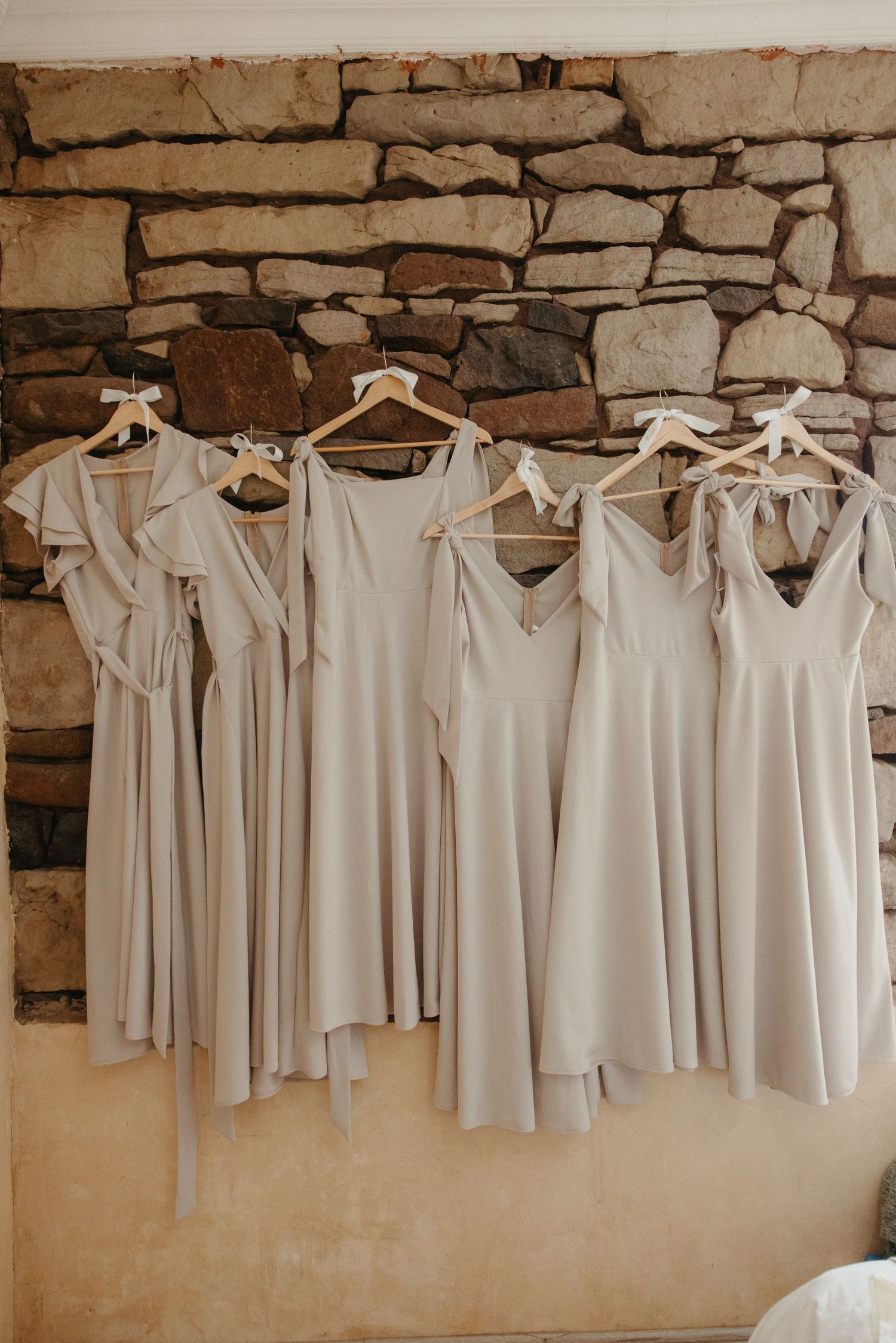 Six mismatched taupe bridesmaid dresses hanging on wooden hangers against a rustic stone wall.