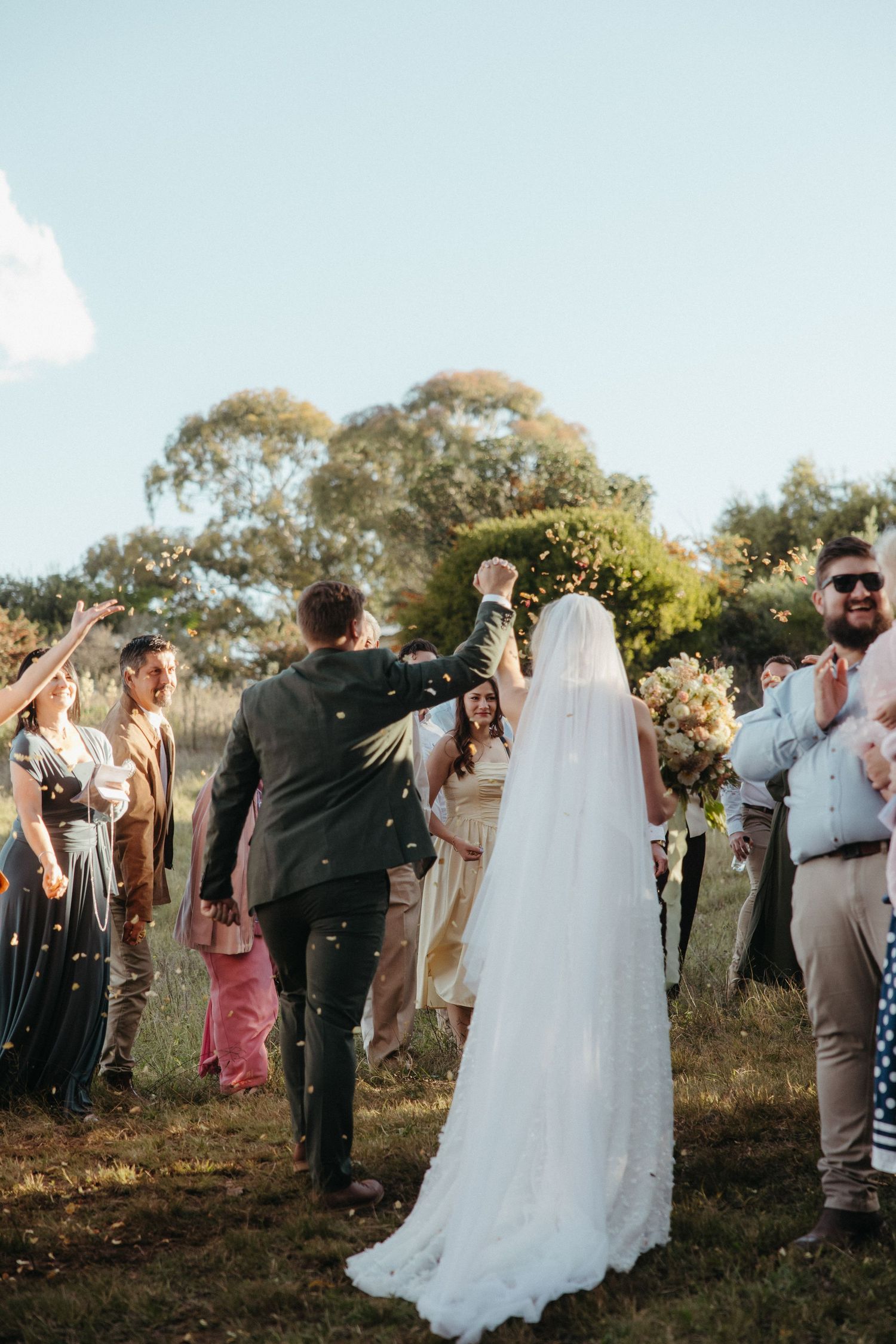 Bride and groom walk through cheering guests tossing confetti outdoors at a sunny wedding ceremony.