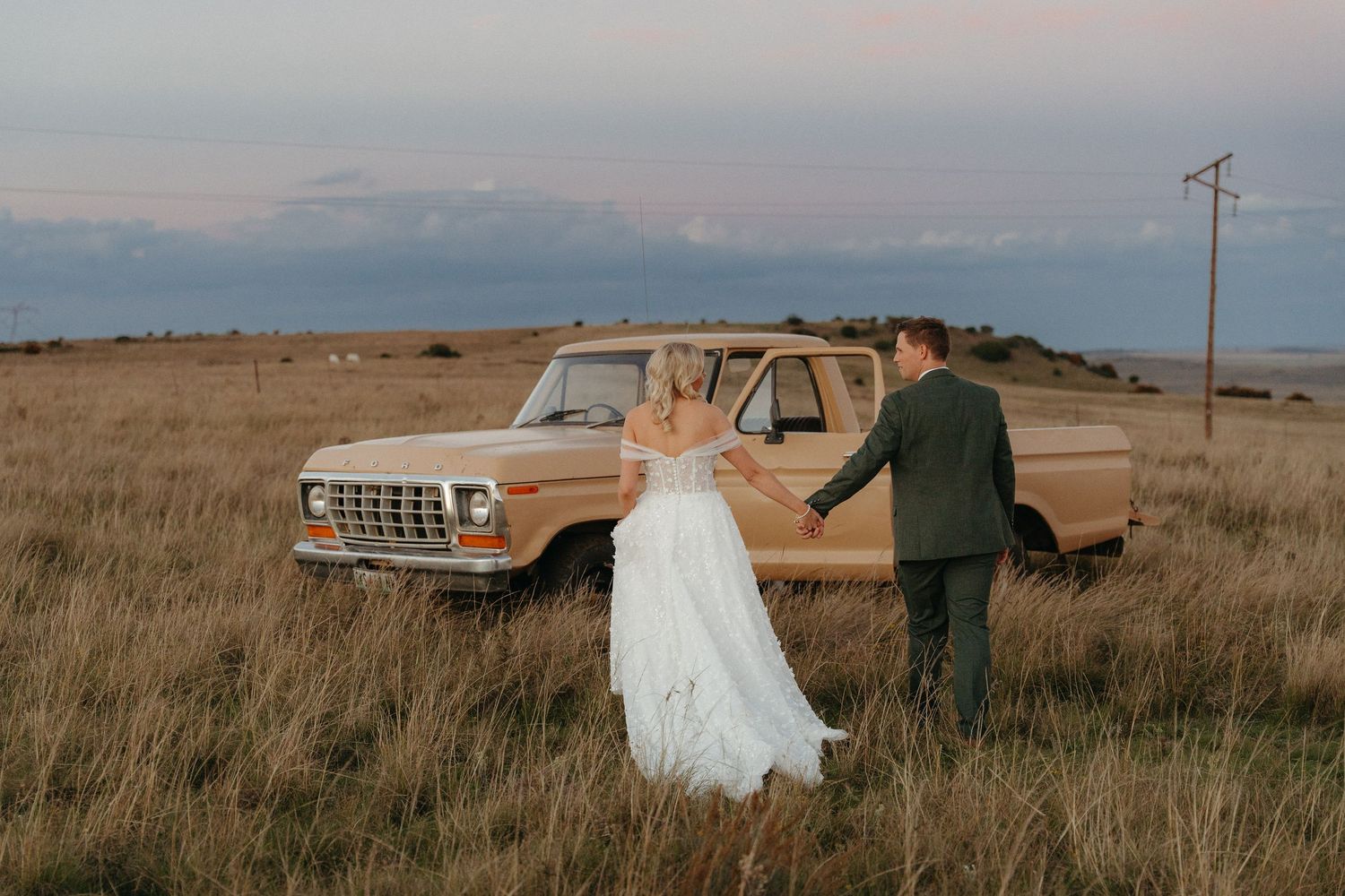 Bride and groom hold hands in golden field at sunset beside a vintage tan Ford truck.