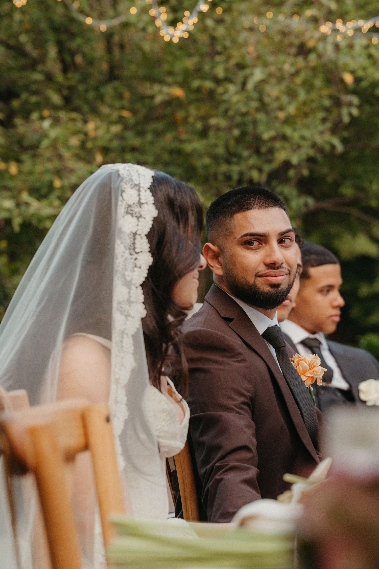 Groom in brown suit smiling during outdoor wedding ceremony, bride in lace veil seated beside him, green trees in background.
