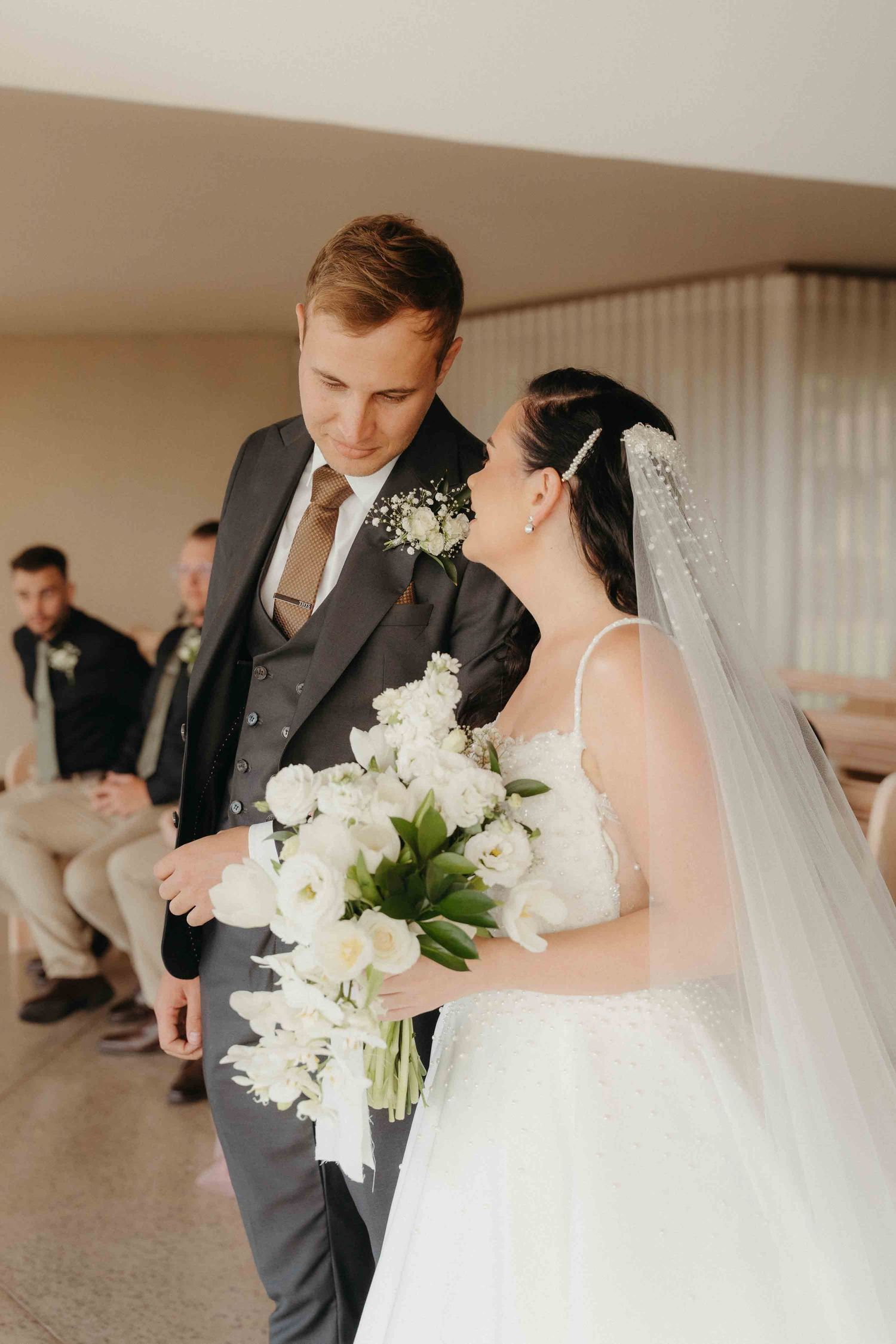 Bride holding white floral bouquet walks down aisle with groom in gray suit during elegant wedding ceremony.