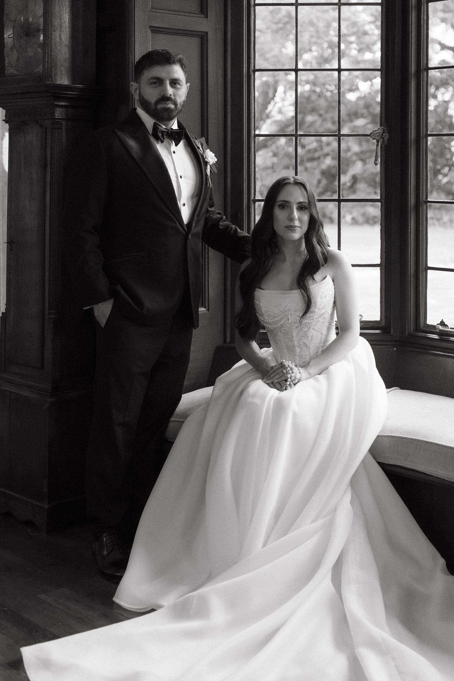 Black and white wedding portrait of bride in strapless gown and groom in tuxedo posing elegantly by a window.