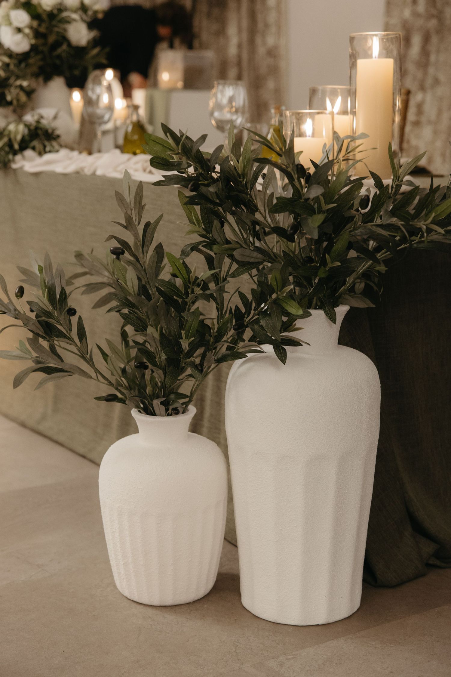 Two white ceramic vases with olive branches beside candles on an elegant table setting.