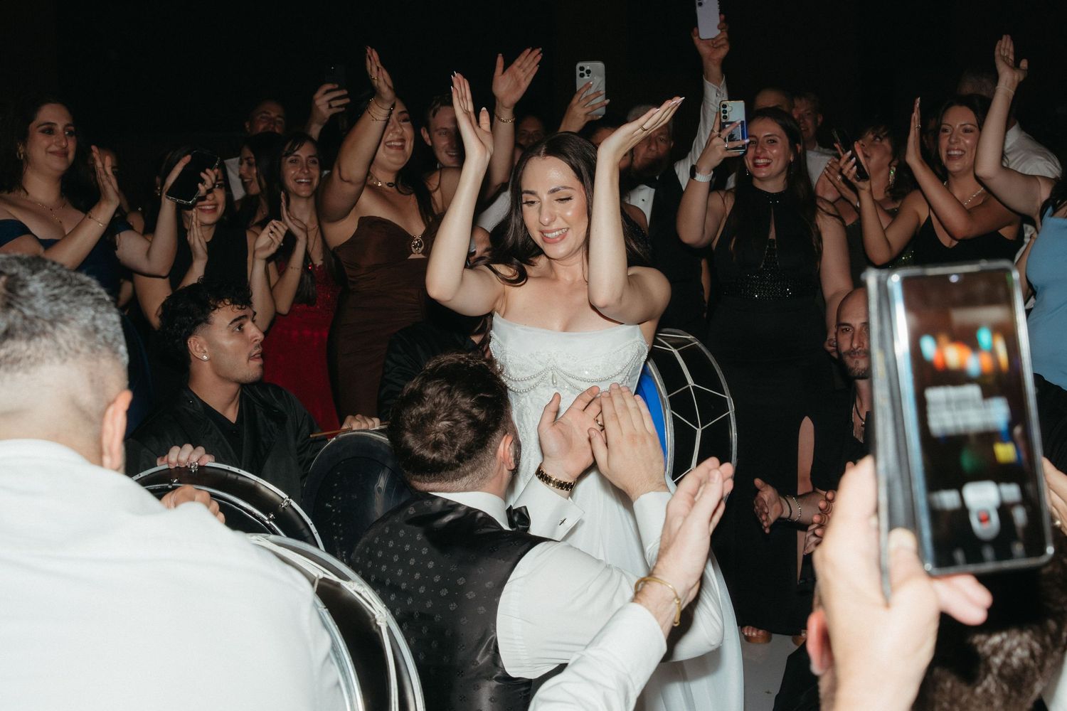 Bride in white dress raises hands joyfully on chair during lively wedding reception dance floor celebration.