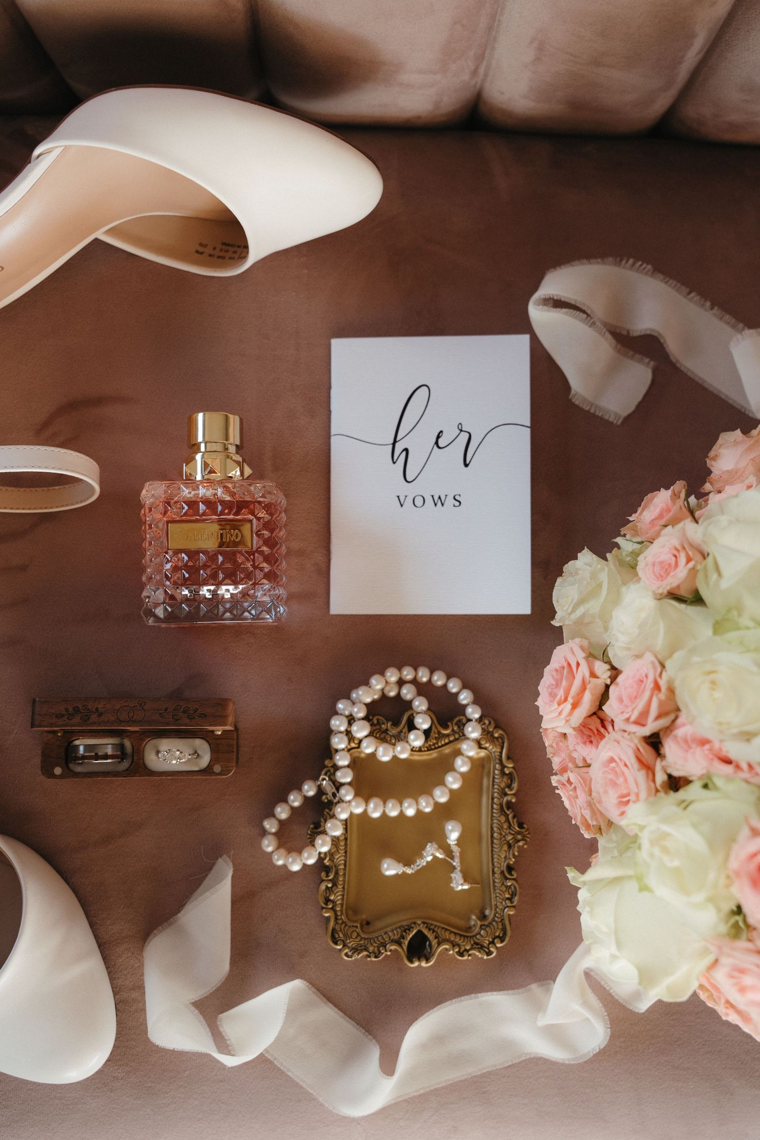 Flat lay of bridal accessories including pearl jewelry, vows card, perfume, rings, and blush heels.