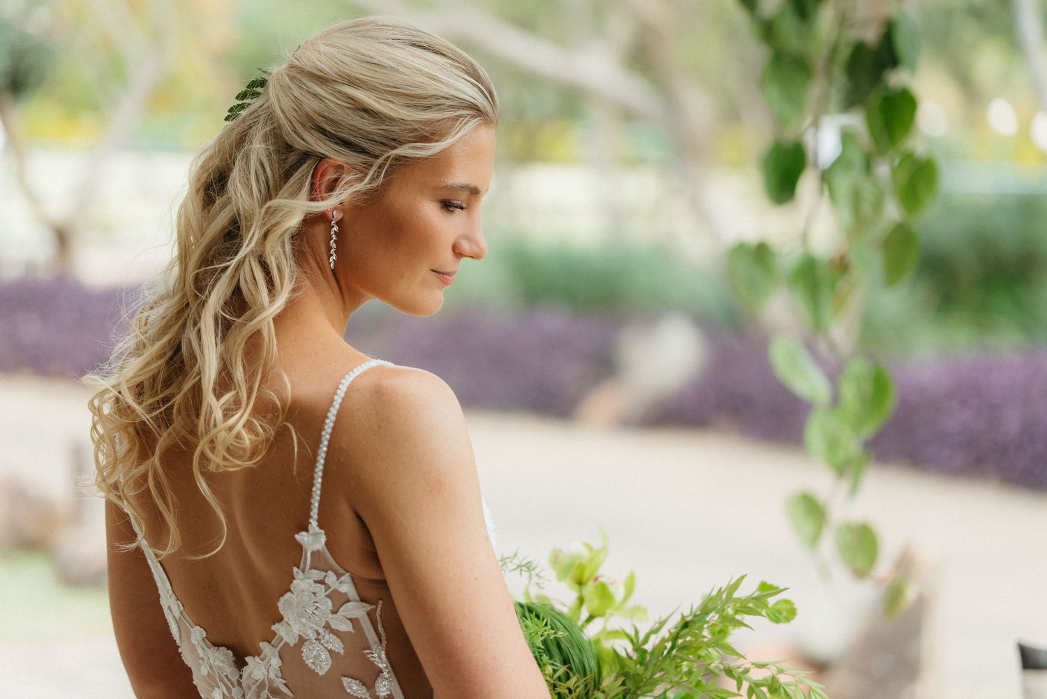 Elegant bride with half-up wavy hairstyle in a backless lace wedding gown holding a green bouquet ar Rosemary Hill, Pretoria