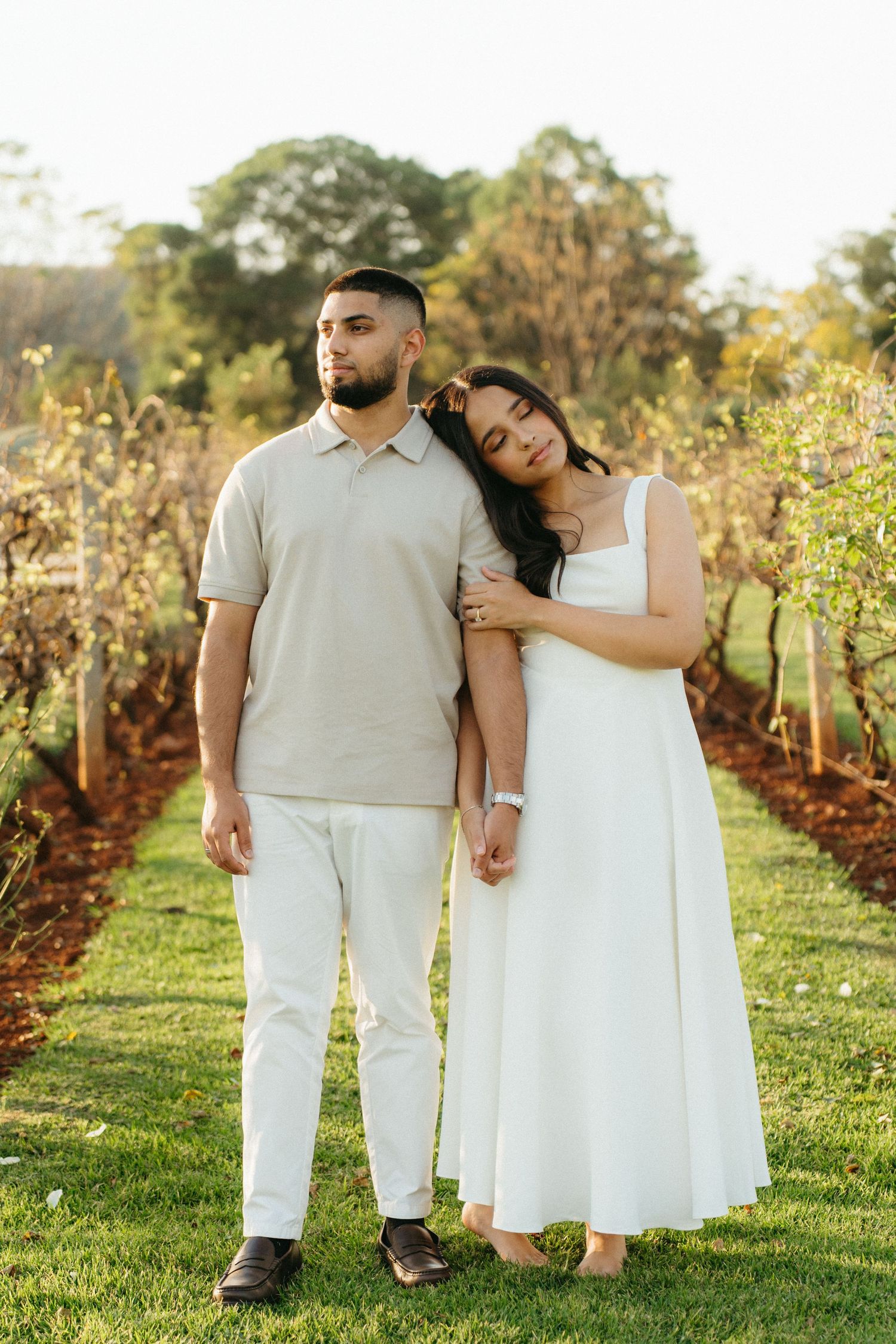 Couple poses in vineyard at Ground the Venue, woman in white dress leans on mans shoulder, golden hour light.