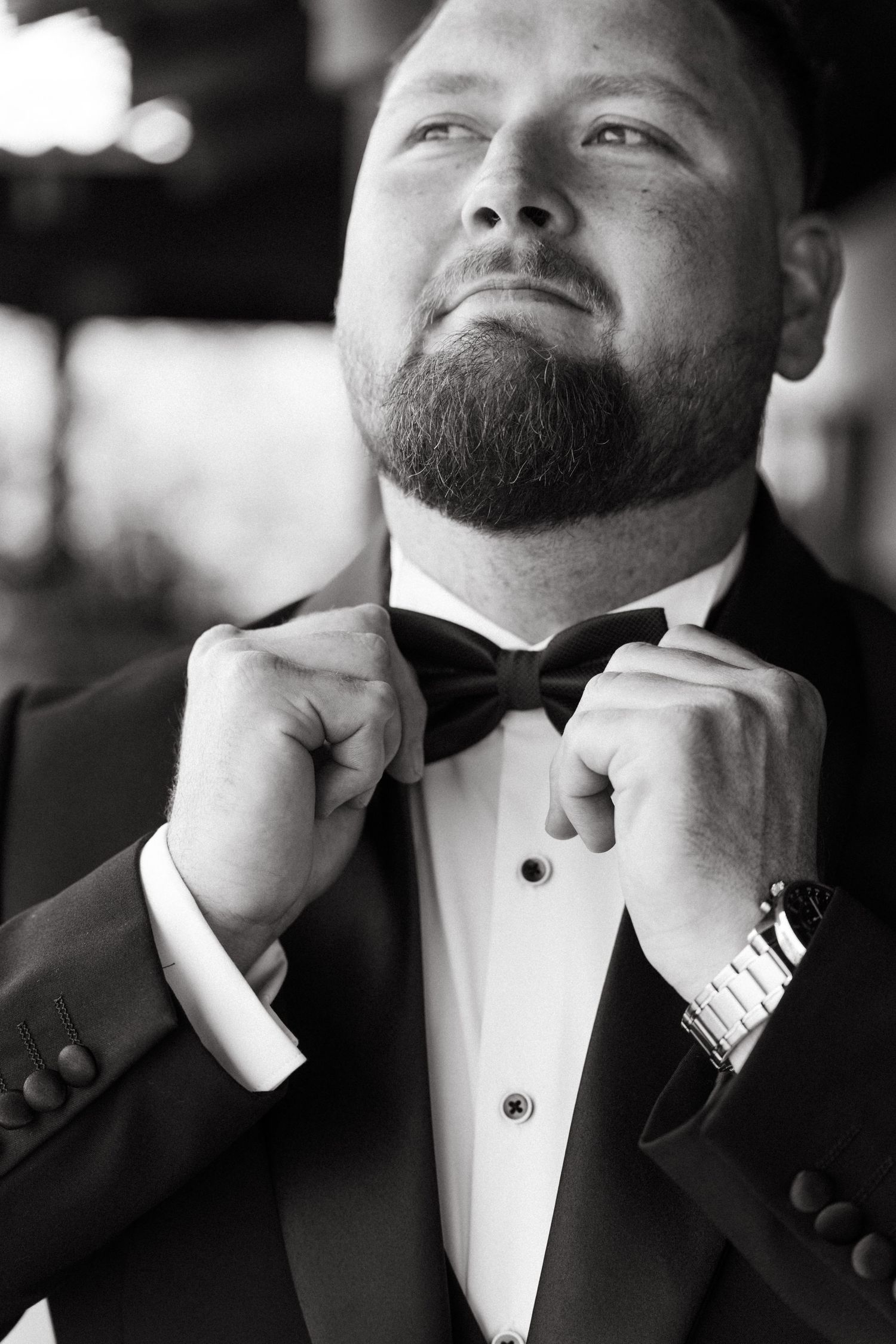 Black and white portrait of bearded groom in tuxedo adjusting bow tie with watch on wrist at Simbali Lodge in Pretoria