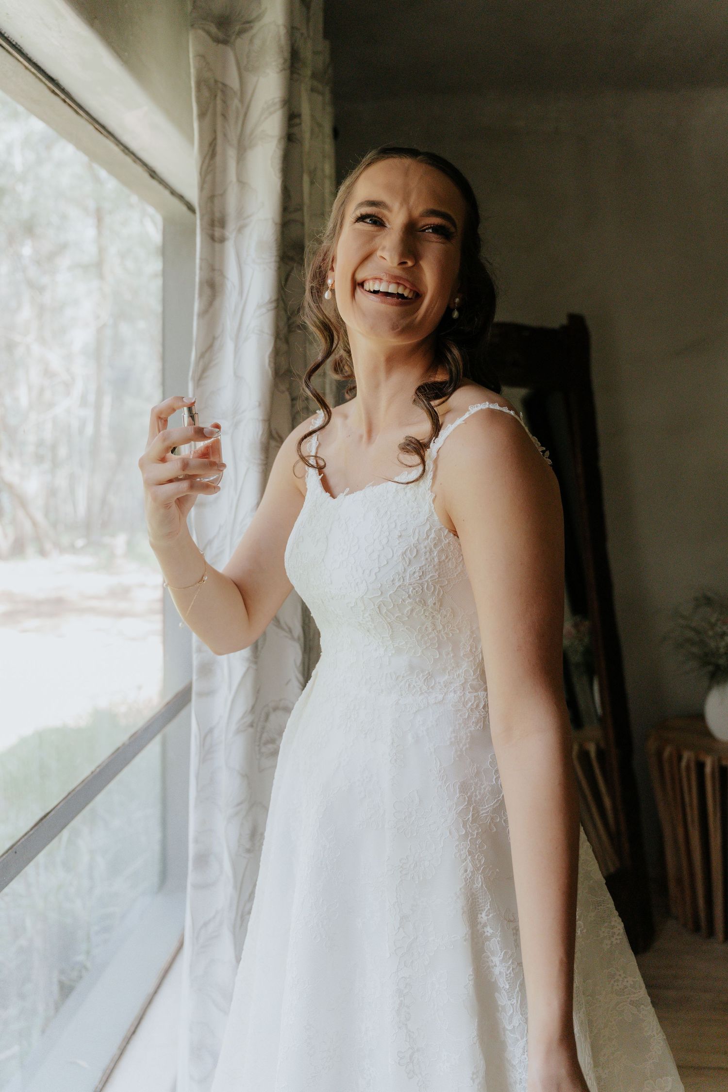A bride in a white lace wedding dress laughs joyfully while standing near a window in natural light.