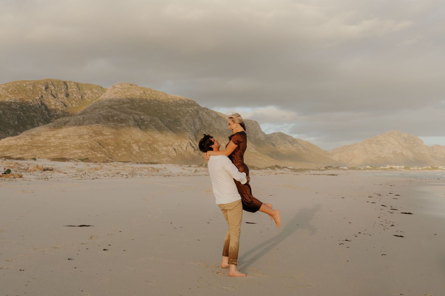 Couple sharing a romantic moment on a sandy beach with dramatic mountain backdrop during golden hour.