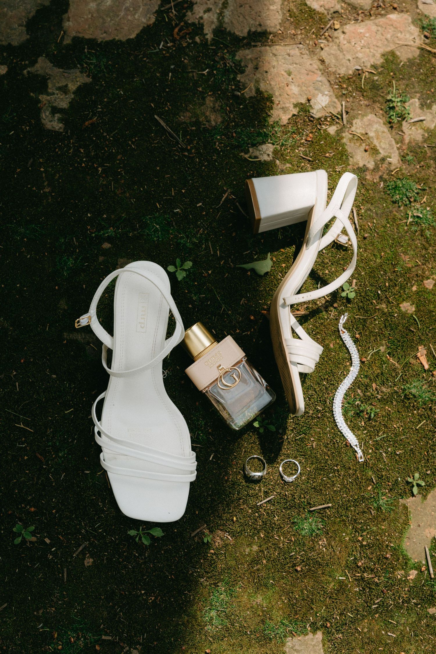 Wedding accessories including white strappy sandals, perfume bottle, and rings arranged on grass and dark background.