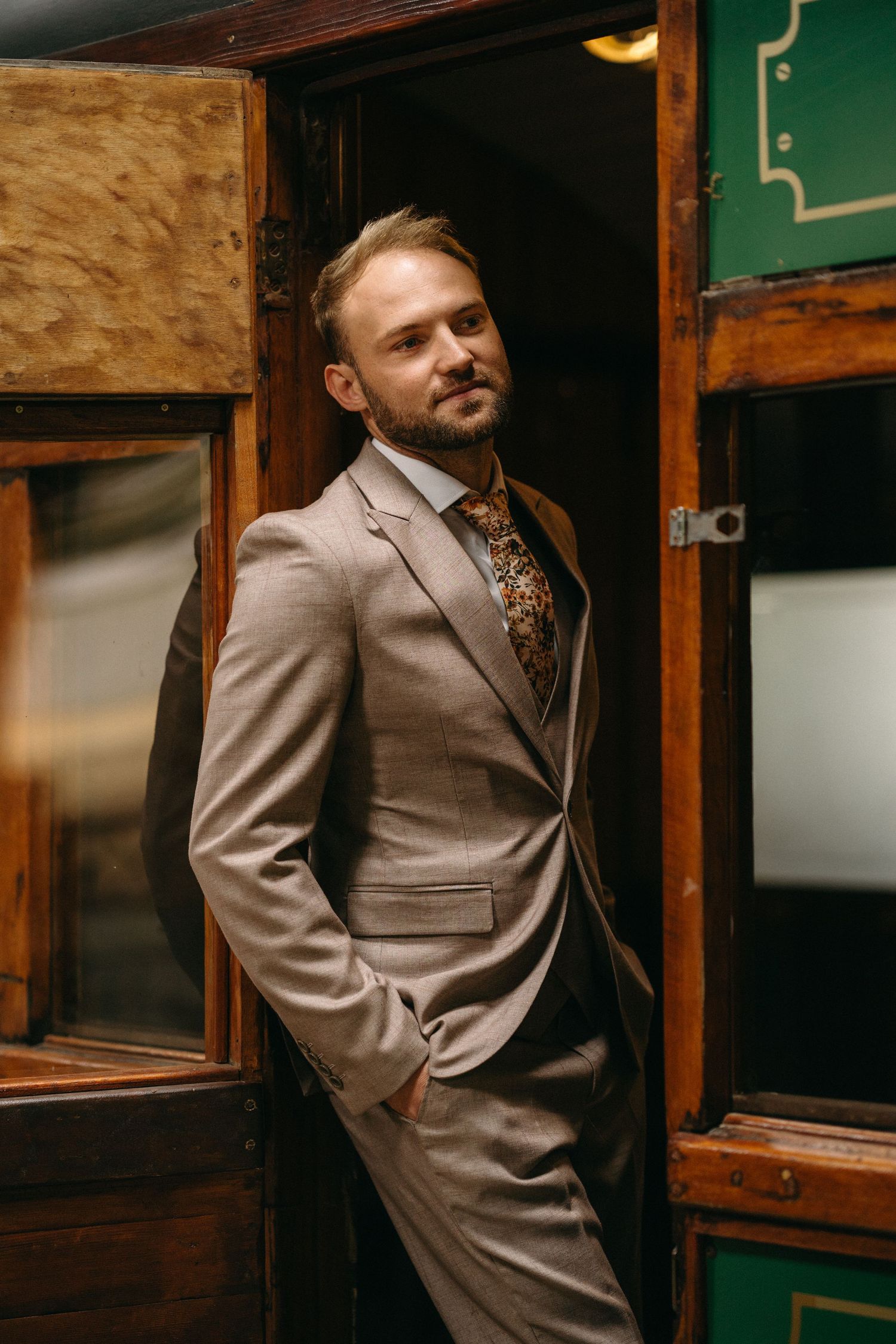 Sophisticated groom in tan suit and floral tie poses in wooden doorframe.
