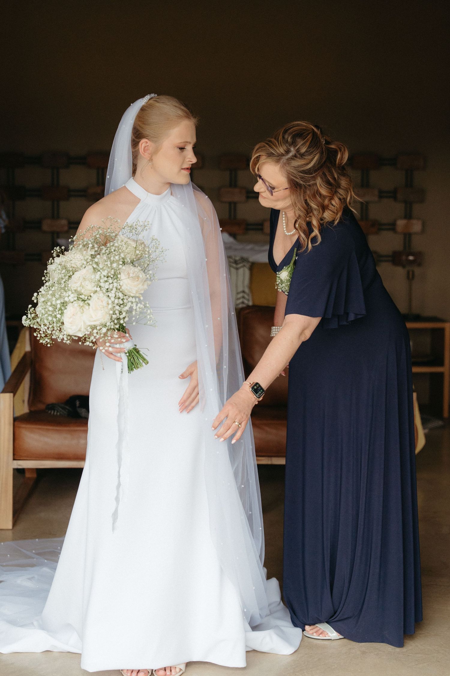 Bridesmaids in navy dresses helping adjust bride's white wedding gown.