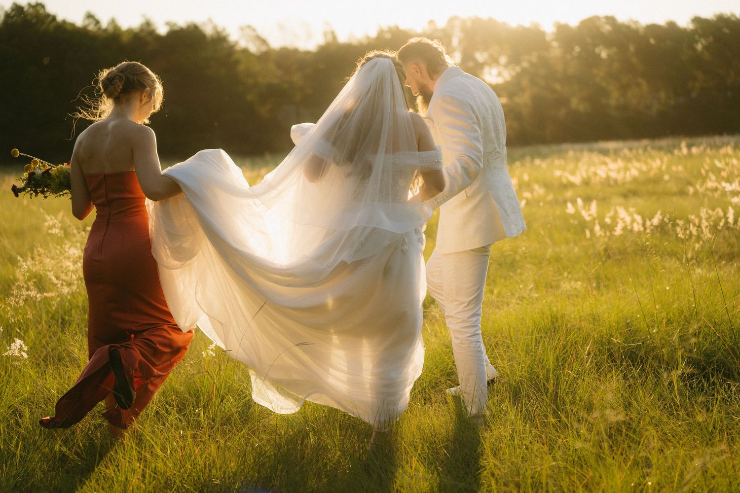 Wedding party helping carry a flowing white veil across a sunlit meadow at golden hour.
