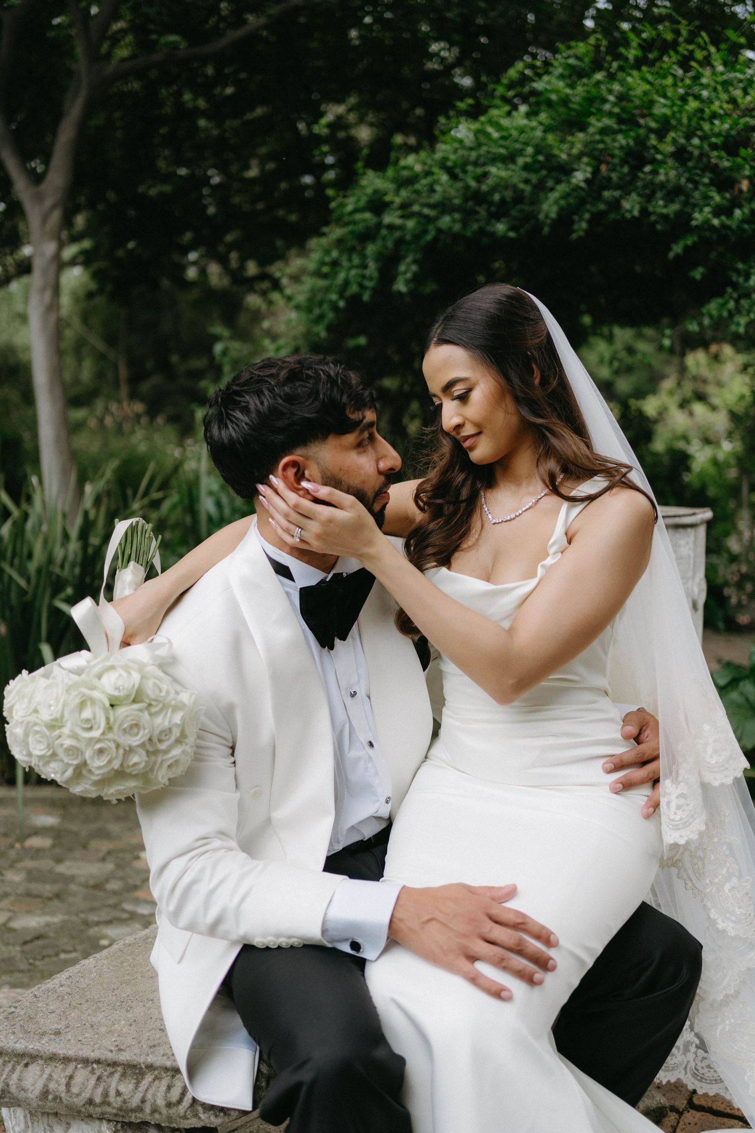 Newlywed couple shares an intimate moment outdoors with the bride in white gown holding a rose bouquet.