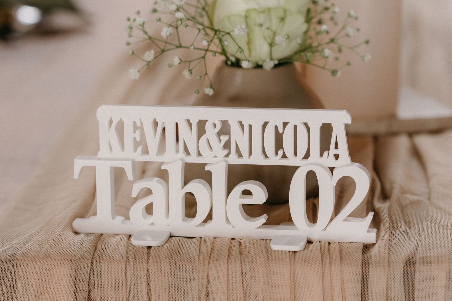 A wooden table number display with white lettering spelling out 'Table 02' with delicate flowers in a vase.