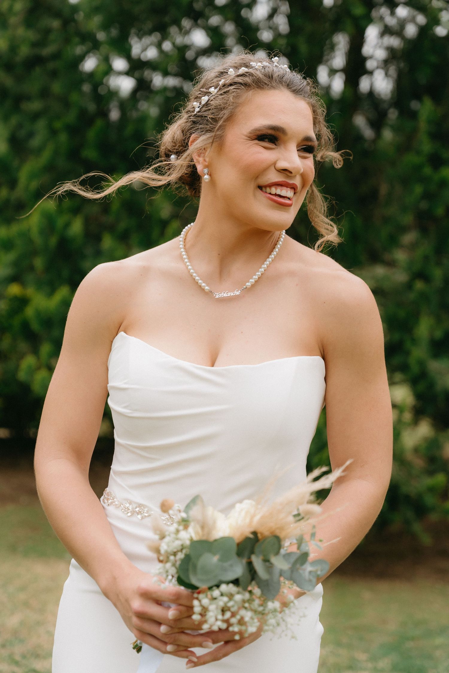 Bride in white strapless gown poses with bouquet against lush green outdoor backdrop.