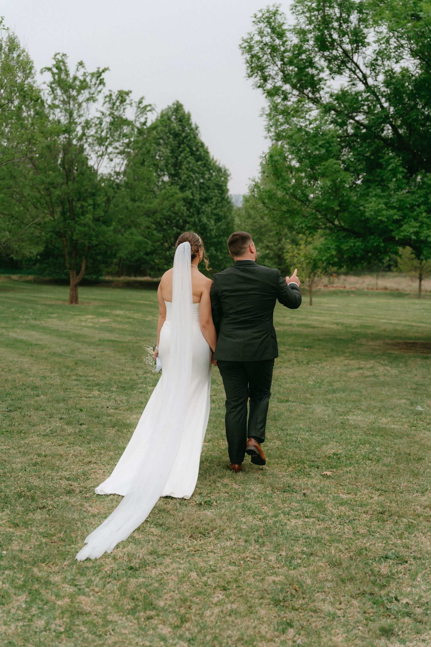 Couple walking across grassy field with bride's veil flowing in the wind.