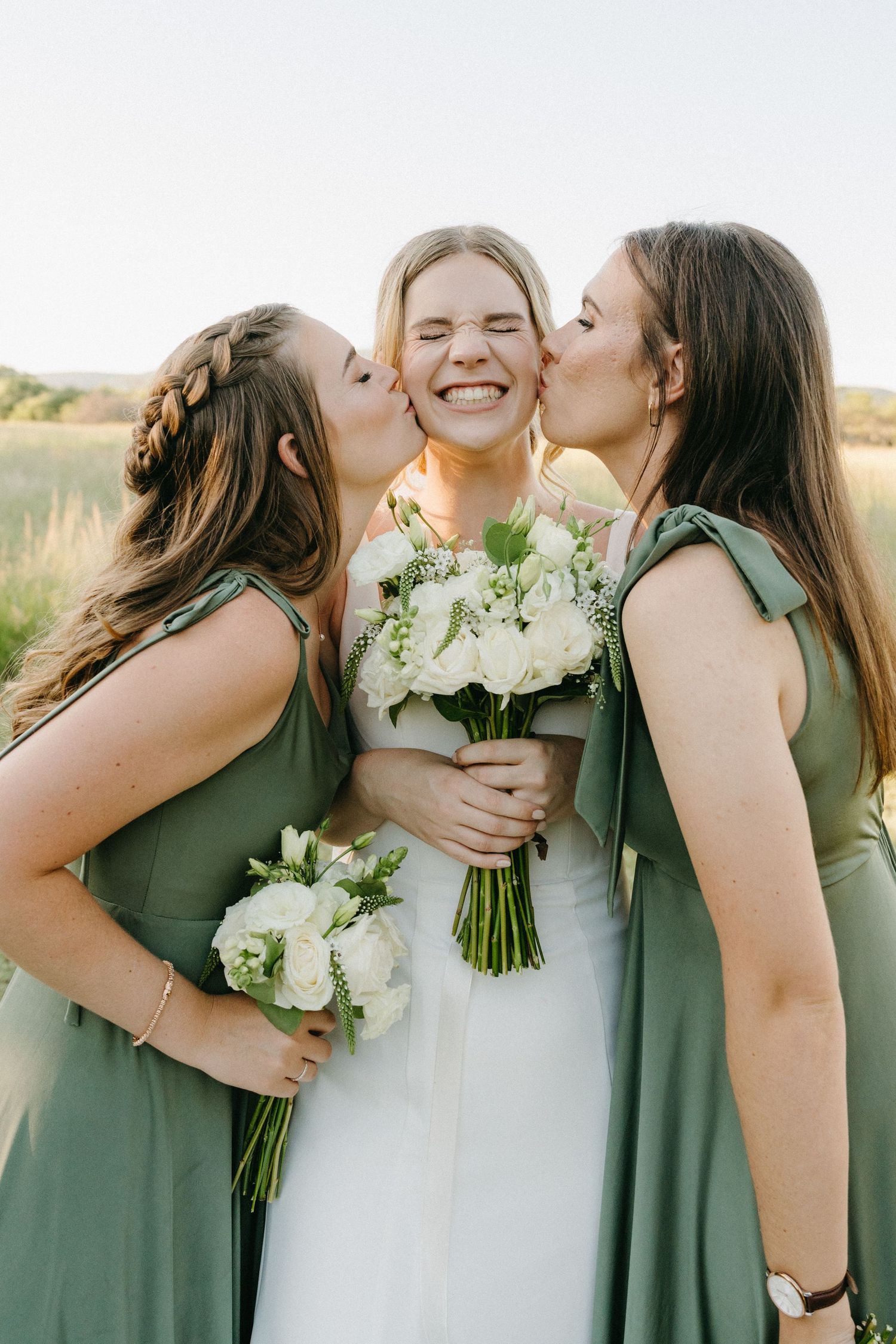 Bridesmaids in sage green dresses kiss bride holding white bouquet during outdoor photo session.