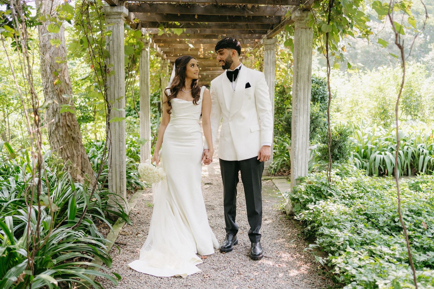 Couple in elegant wedding attire walking hand in hand under a lush garden pergola covered in greenery.