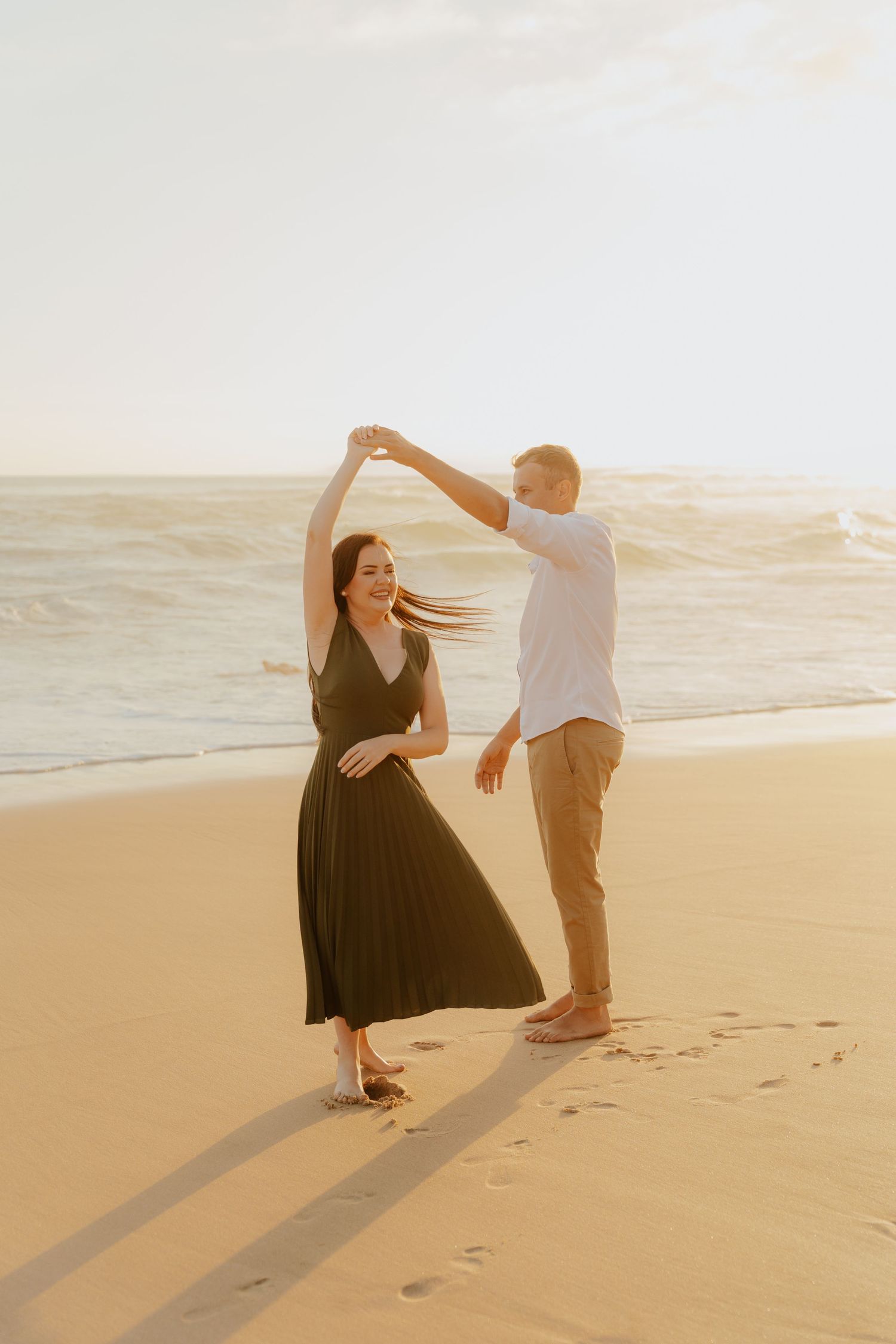 A couple dances together on a sunlit beach during golden hour, casting long shadows in the sand.
