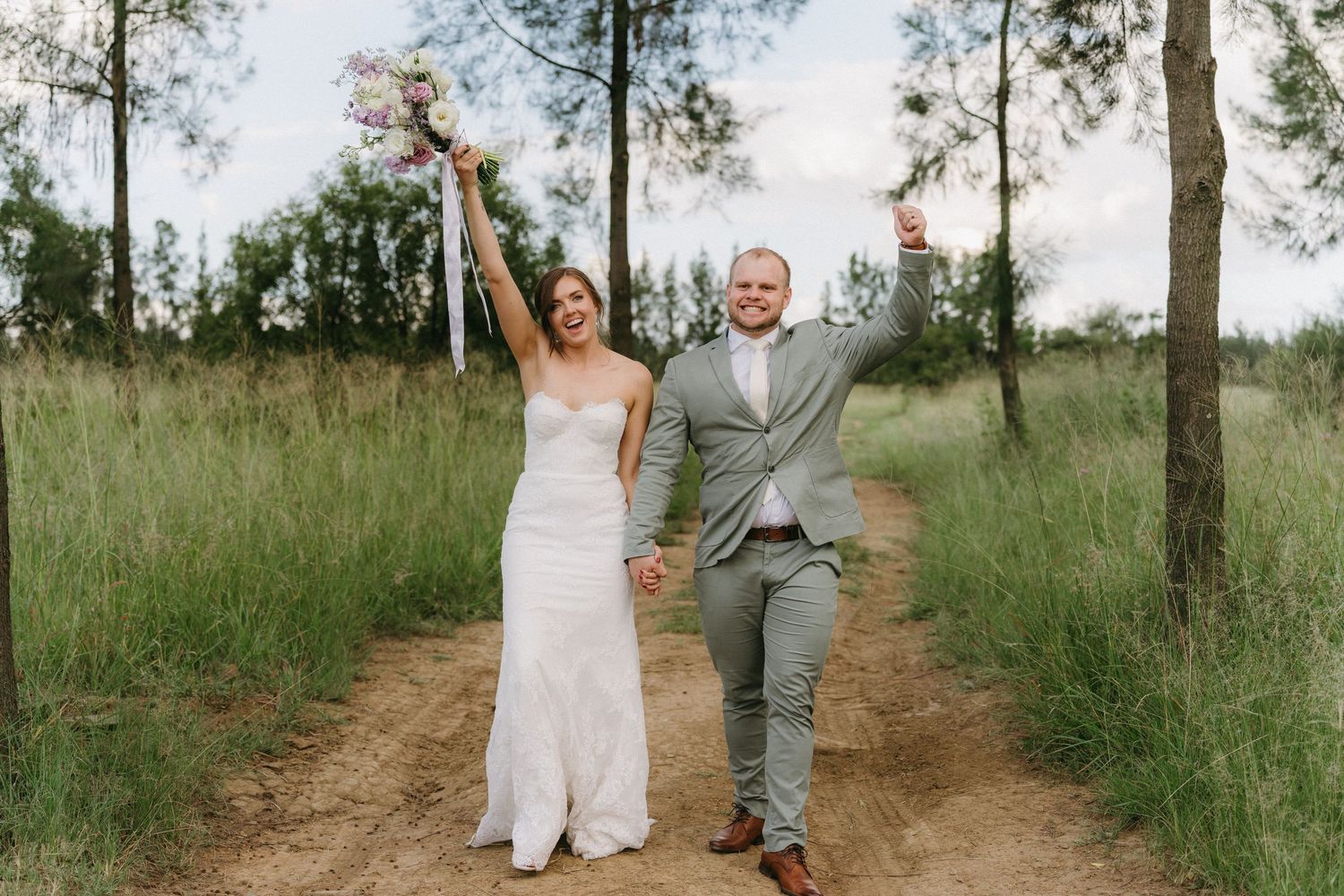 A joyful celebration on a dirt path through tall grass as the newlyweds raise their hands in triumph.
