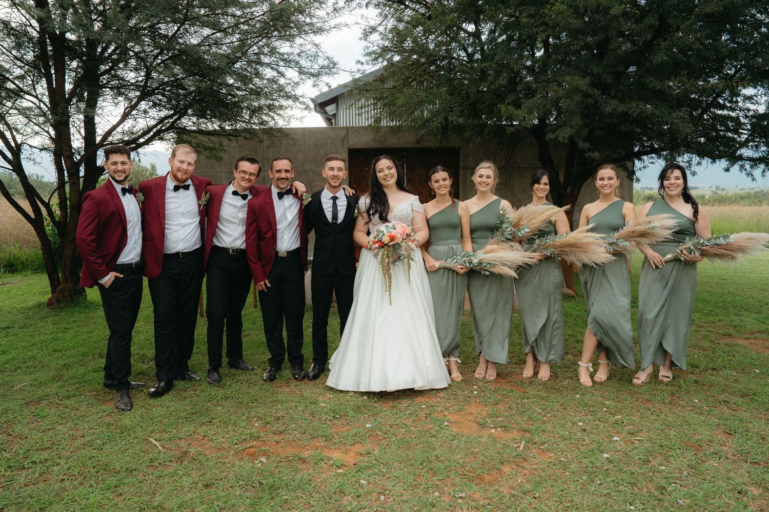 Wedding party poses under a large tree in outdoor setting wearing grey and burgundy attire.