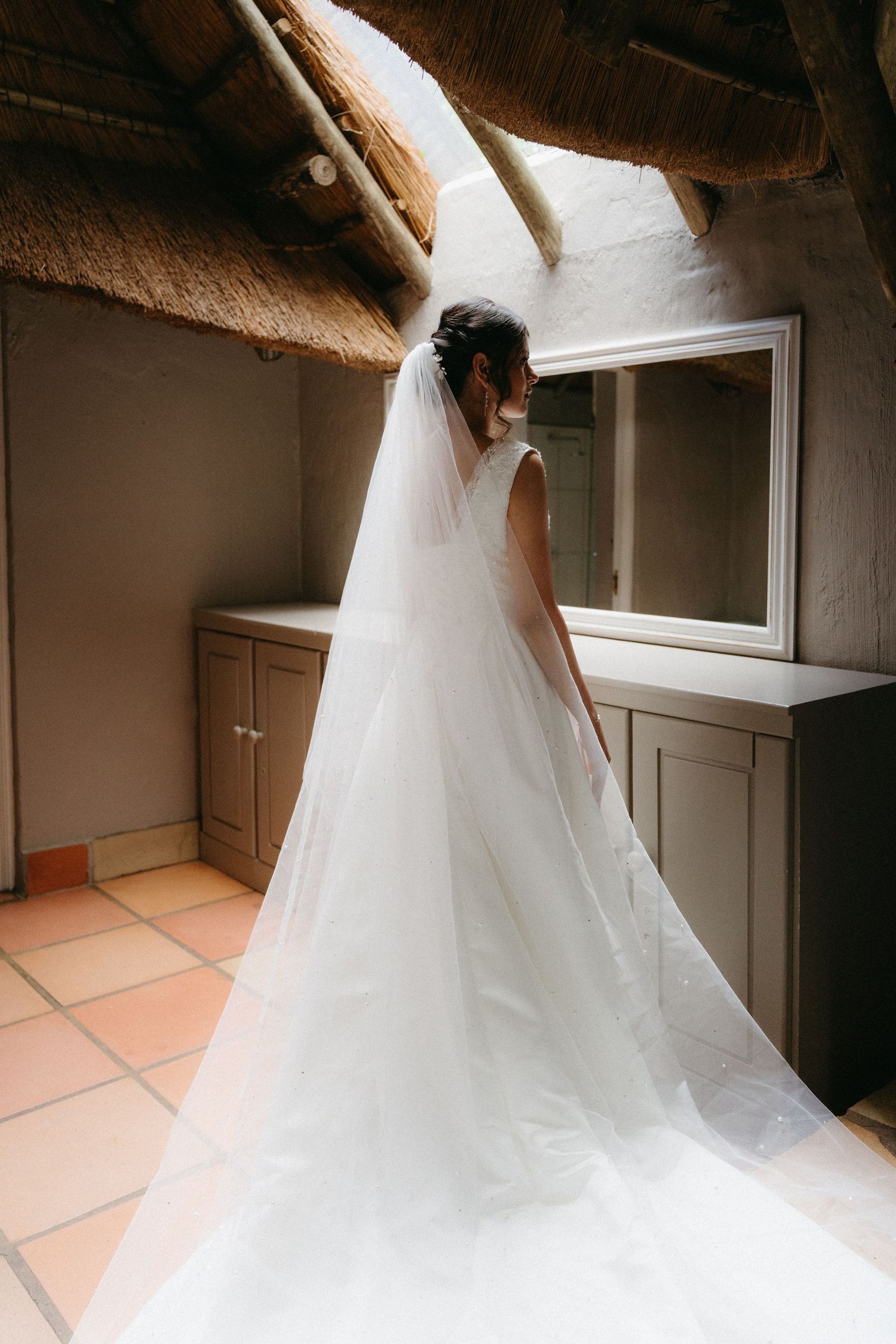 A flowing white wedding gown with a long train and veil photographed in a rustic room with terracotta floor tiles.