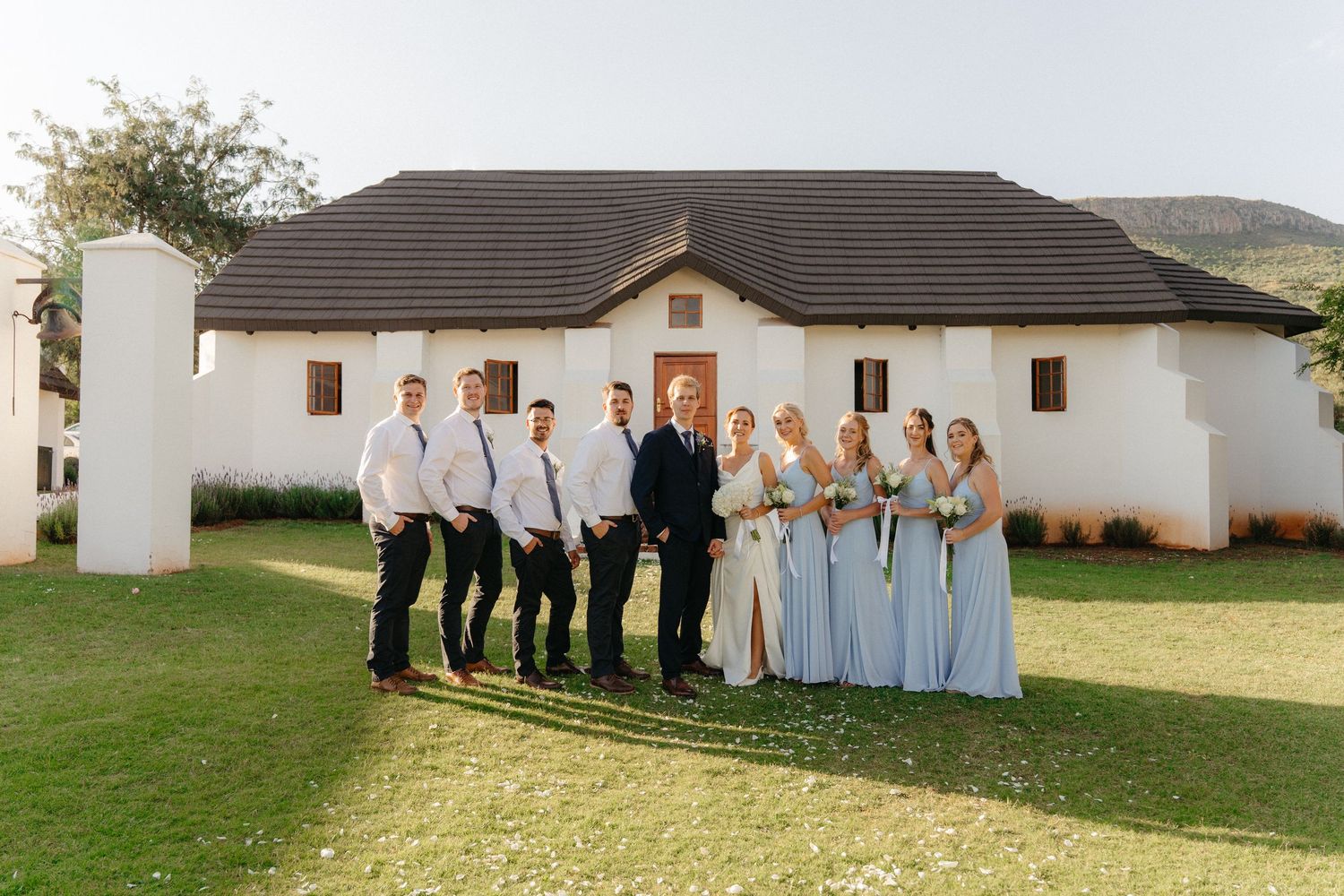 Wedding party members stand in a row on grass outside white buildings in coordinating formal attire.